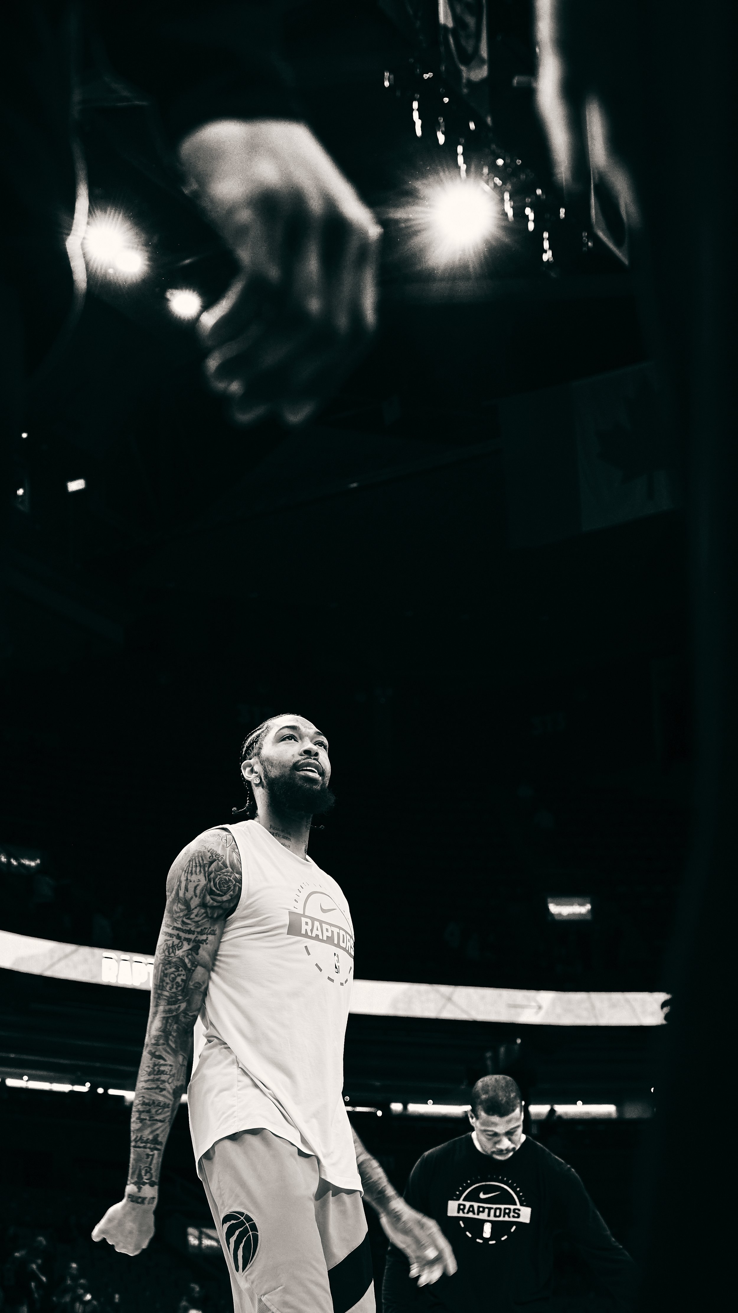 A basketball player with tattoos on his arms wearing a Toronto Raptors uniform standing on a basketball court during a game, looking upward. Another person wearing a Toronto Raptors hoodie is nearby, with a dark background and bright lights overhead.