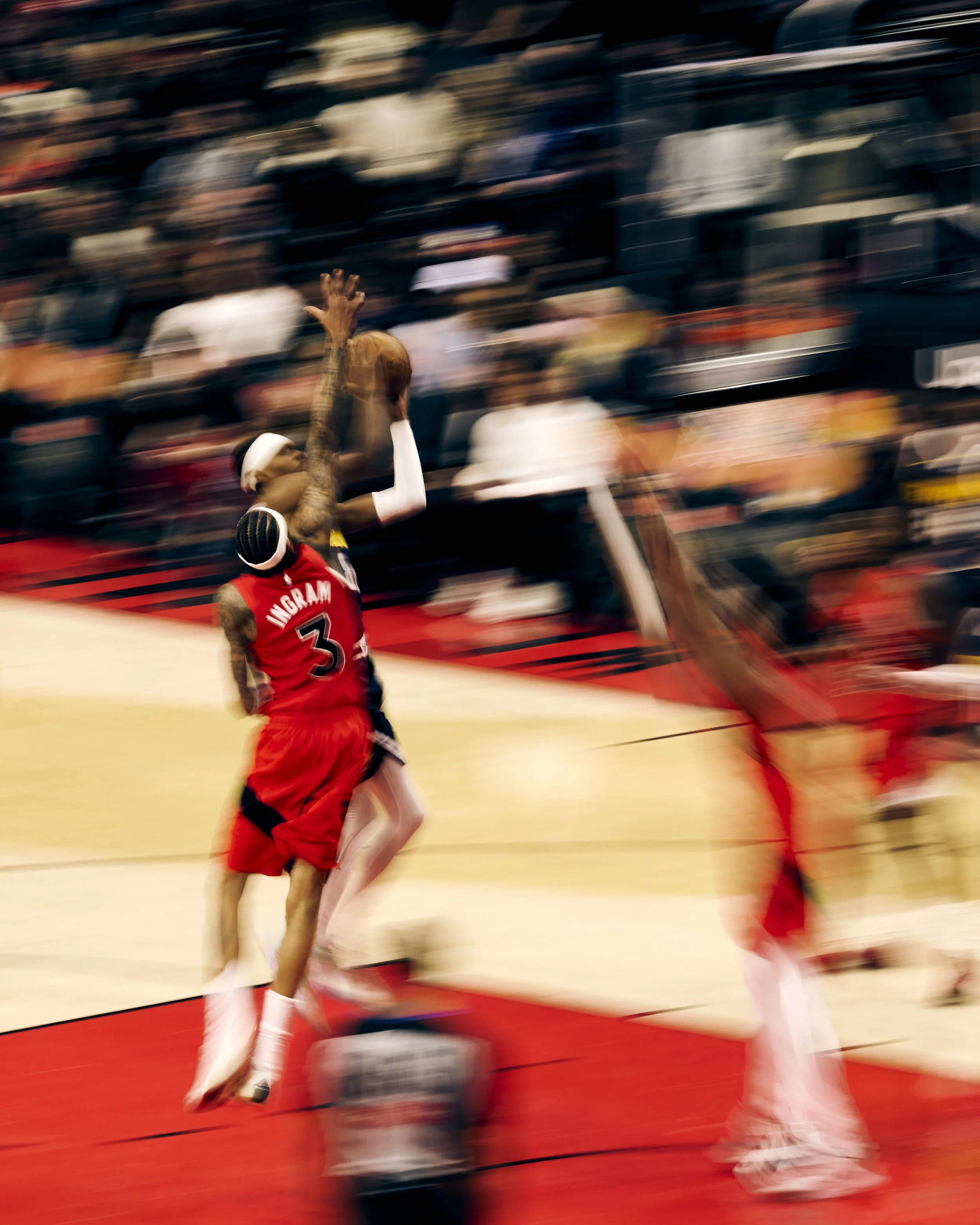 A basketball player wearing a red jersey with the name 'Ingram' and the number 3 is jumping to make a shot or dunk during a game. The background shows a blurred crowd in the stands, emphasizing the motion and action of the scene.