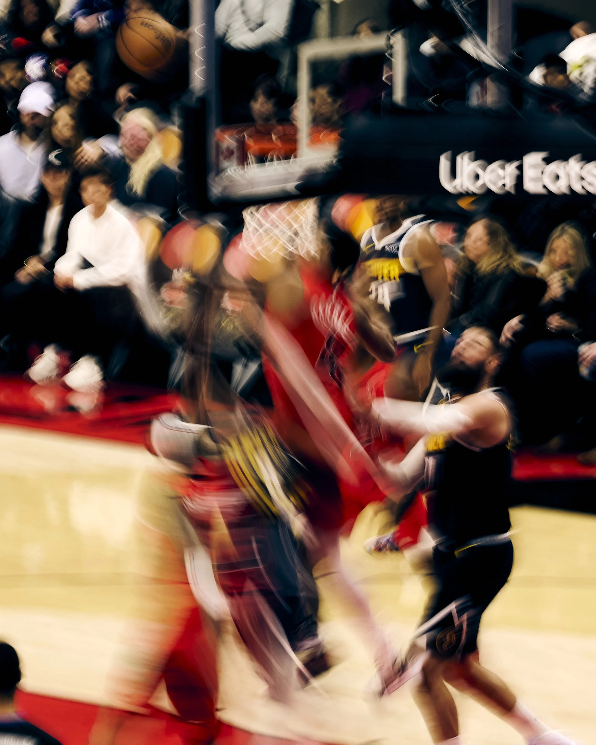 Blurred action shot of a basketball game with players, spectators, and a basketball hoop visible.