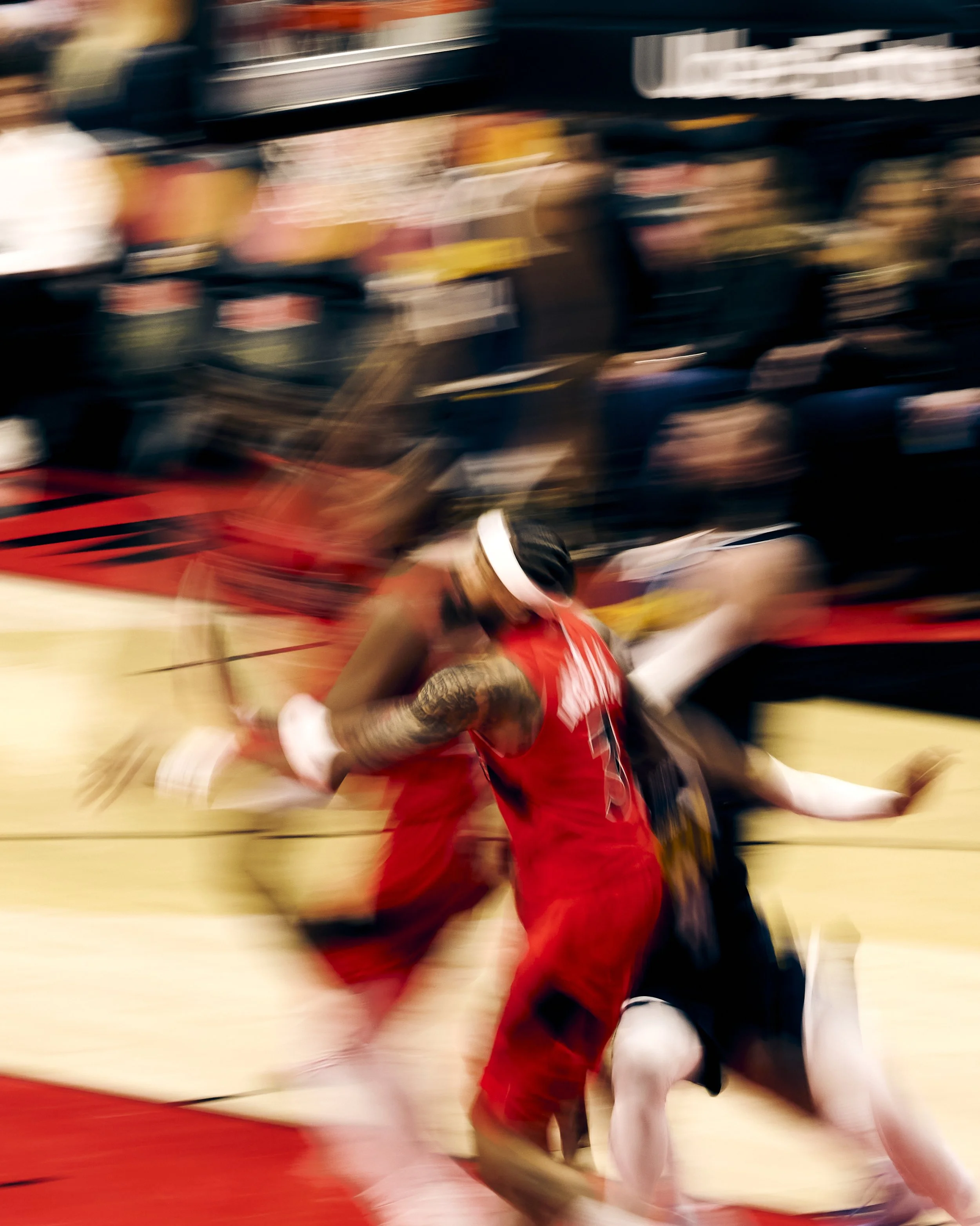 Blurred action shot of two basketball players in red and black uniforms competing on a court.