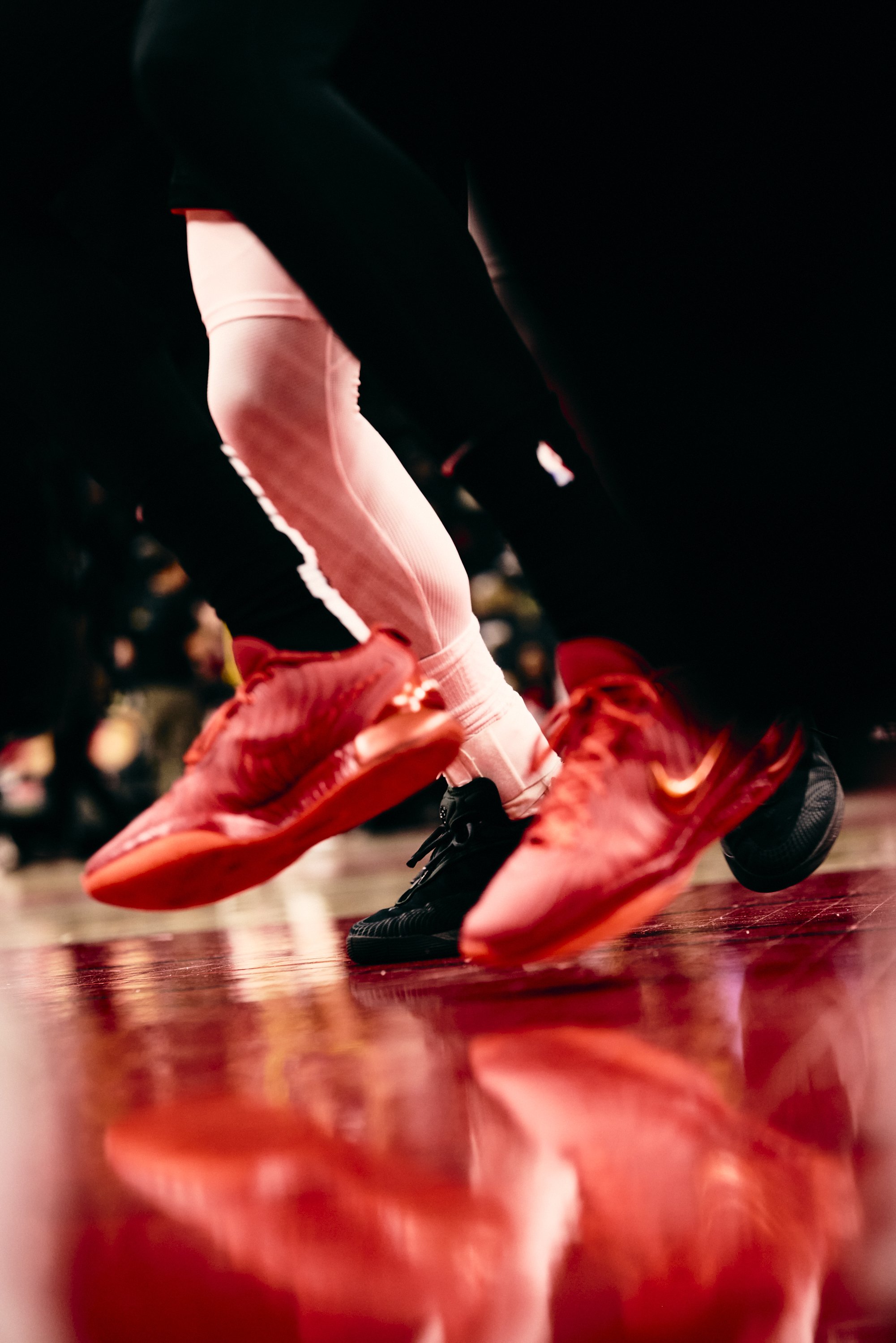 Close-up of a person in red sports shoes and white athletic pants, bending down on a wooden floor during a sports event.
