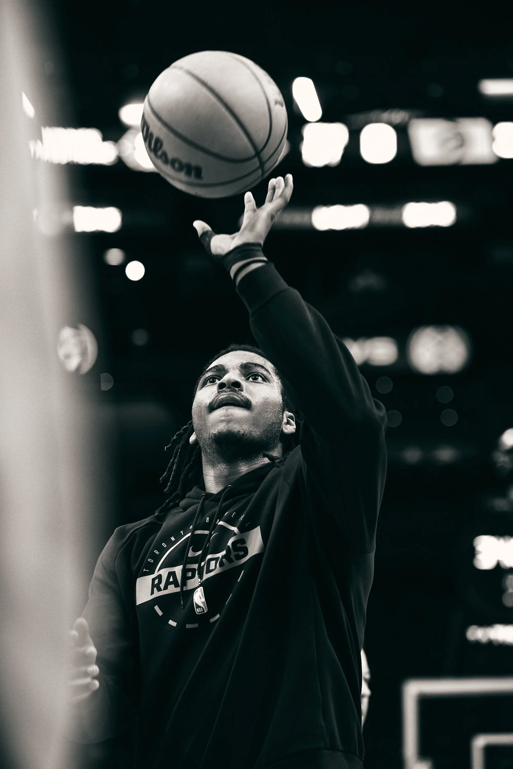 A basketball player taking a shot, wearing a Toronto Raptors hoodie, in a dark indoor stadium with blurred lights in the background.