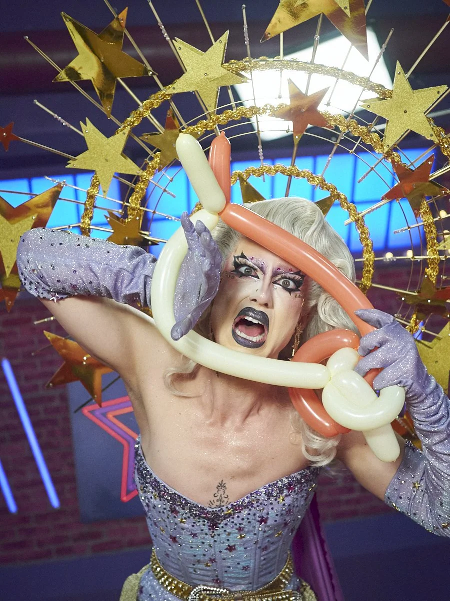 Saltina Shaker a Drag Queen on Canada's Drag Race Season 6, with platinum blonde hair styled in curls, wearing a sparkling purple strapless dress and glittery purple gloves.