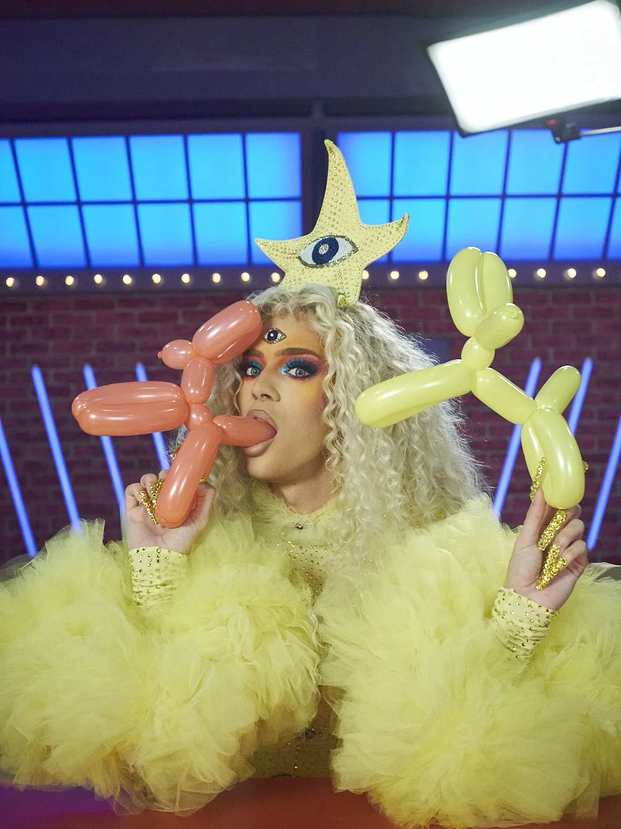 Paolo Perfección with curly platinum blonde hair in yellow fluffy outfit and eye decorations, holding balloon animals, with a star and eye headpiece, in a colourful, neon-lit setting. Season 6 contestant of Canada's Drag Race.