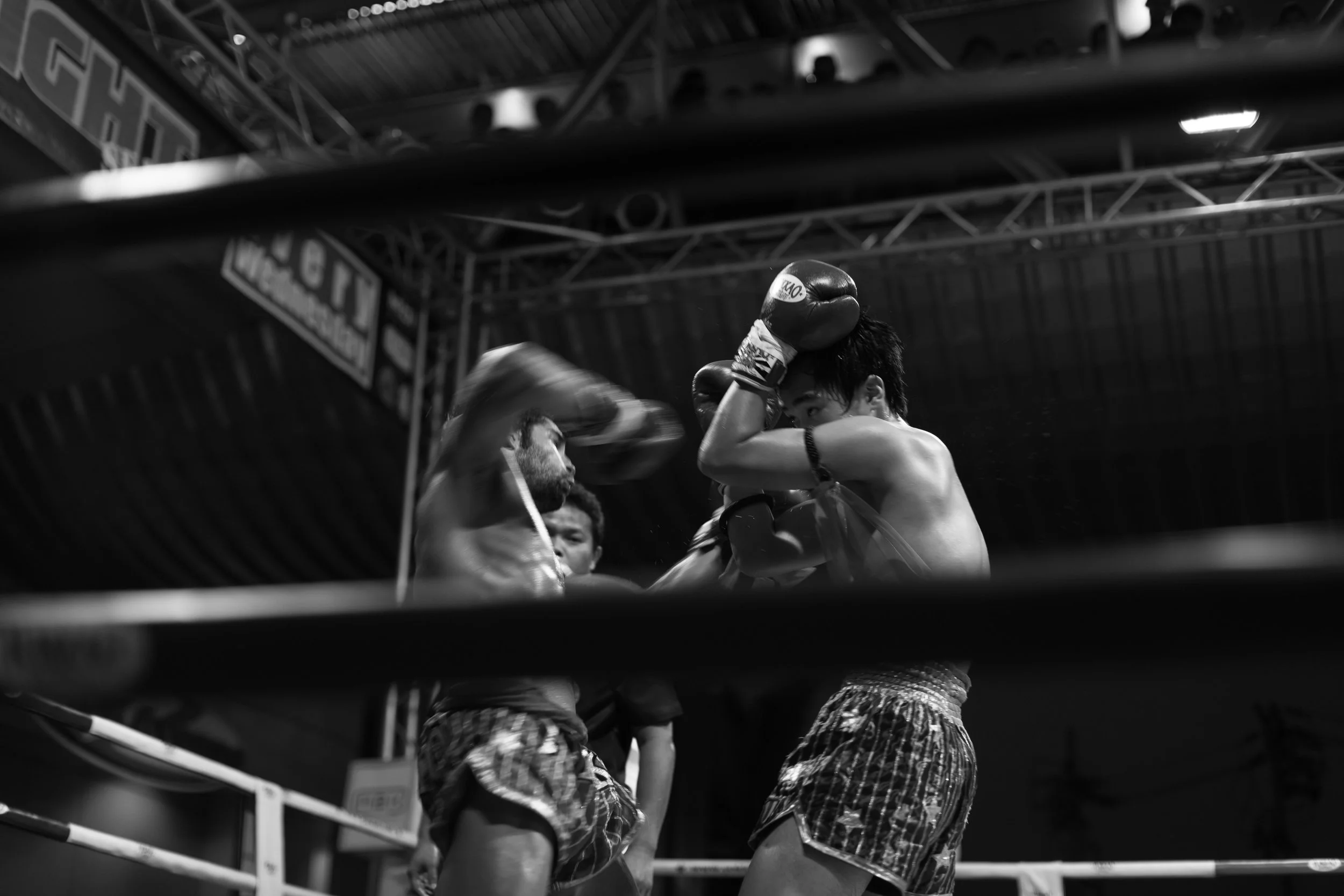 Thailand, Muay Thai, black-and-white photo of two young male boxers sparring in a ring, wearing boxing gloves and shorts. One boxer's face is partially visible, and he is throwing a punch towards the other. Ring side during a Muay Thai match in Thail