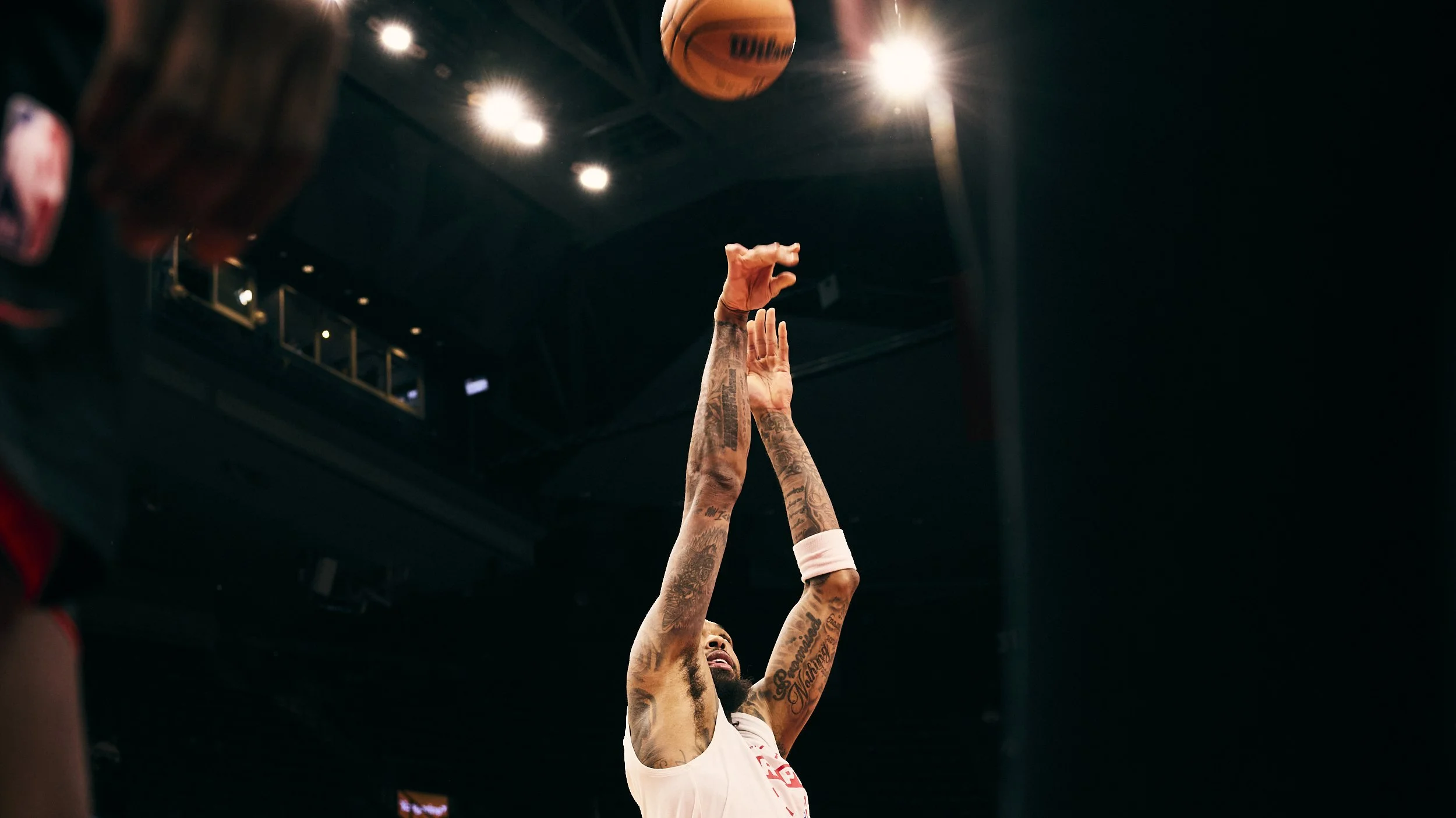 A basketball player making a shot during a game, with his arm extended upwards and the basketball leaving his hand, against a dark background with bright overhead lights.