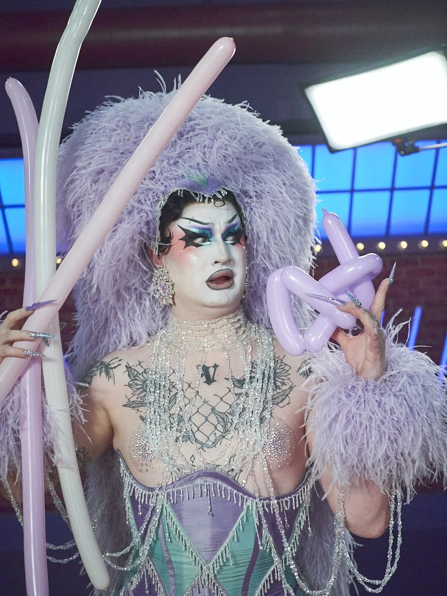Van Goth, Winner of season 6 of Canada's Drag Race, wearing an elaborate purple costume with a feathered headdress, dramatic makeup, and rhinestone jewelry, holding balloon sculptures and sticks.