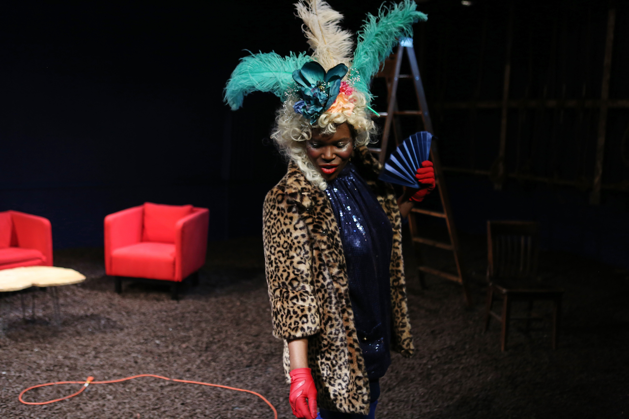  Julienne "Mizz June" Brown as Marsha P. Johnson in the foreground looking down wearing flowers and feathers on her head and a faux-fur leopard-print coat and red leather gloves. She holds a fan in her right hand. Dirt covers the floor the red chairs