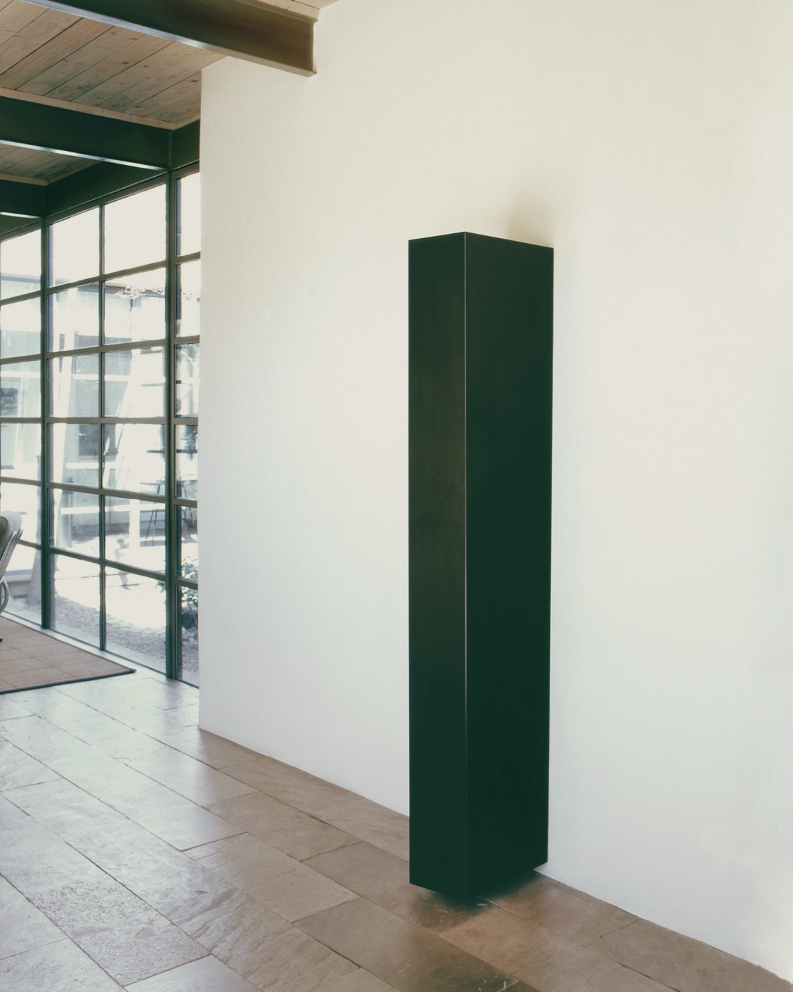 Long rectangle made of graphite, attached to a white wall, but floating just above the floor.