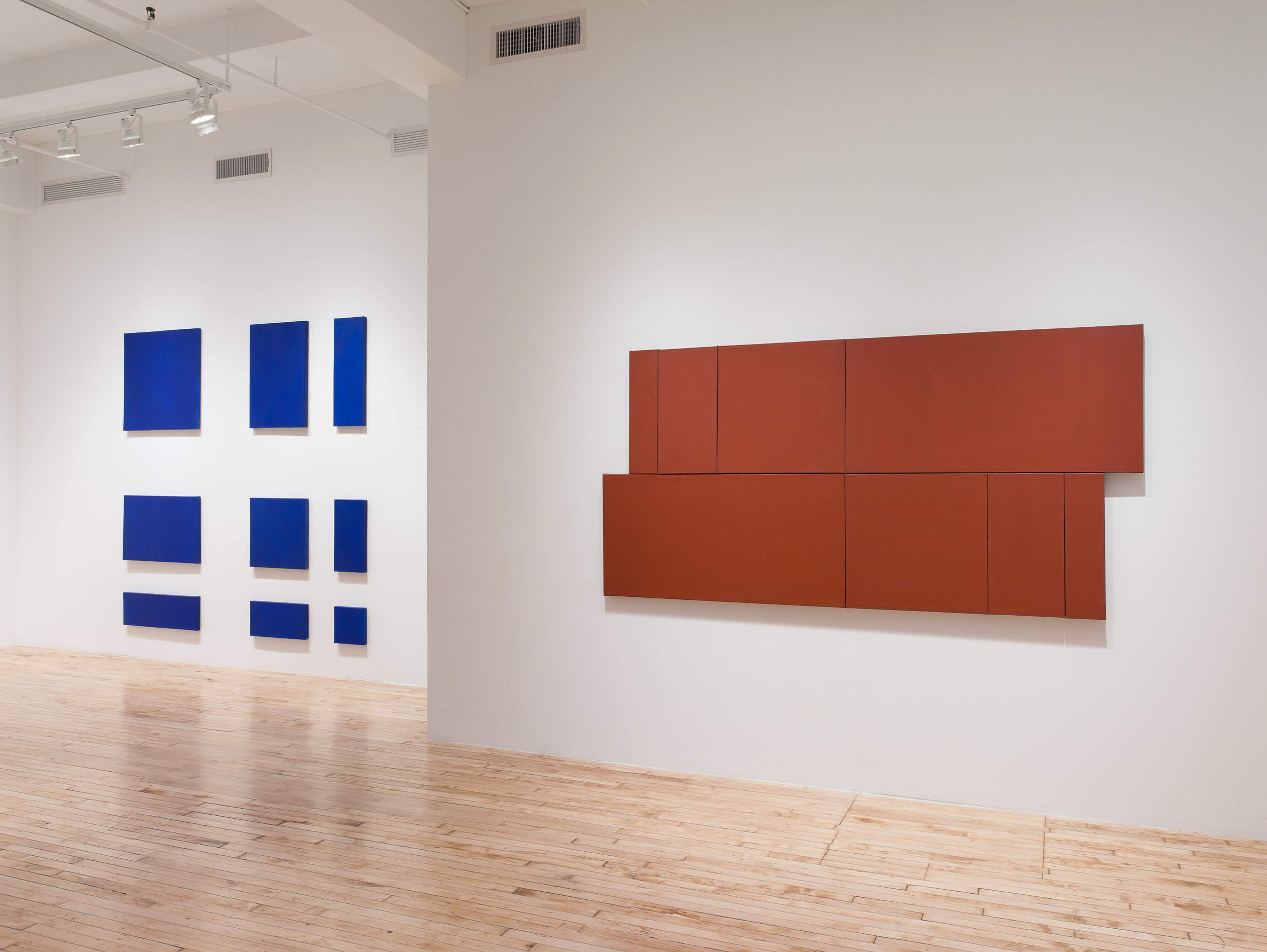 Grid of blue rectangles on the far wall of the gallery. Long rust colored canvases hang on the wall in the foreground.