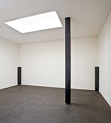 Three floating columns of graphite in a white room. The two in the corners are half the height of the wall and the one in the center hangs from the ceiling to just above the floor.