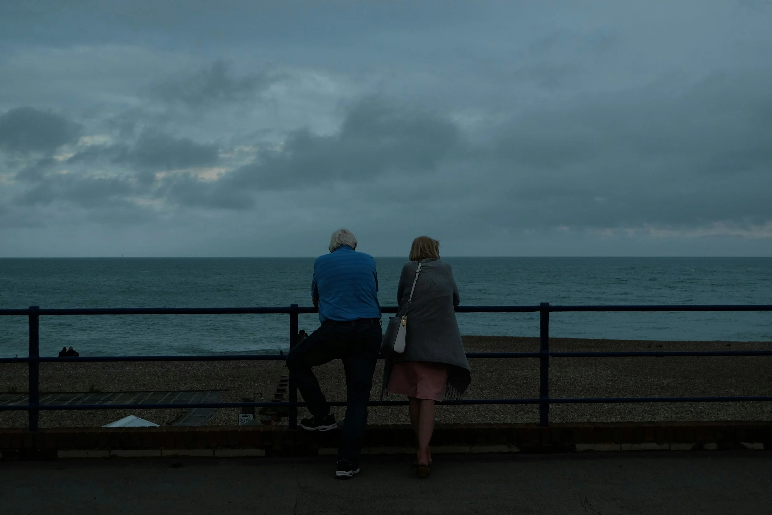 Two people sitting on a bench by the ocean, facing the water, under a cloudy sky.