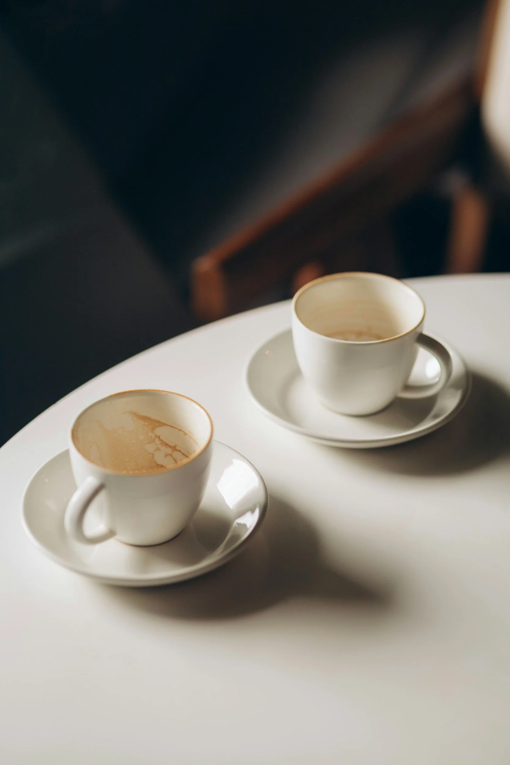 Two white ceramic coffee cups on matching saucers placed on a round white table. The cups contain coffee with remnants of coffee on the inside.