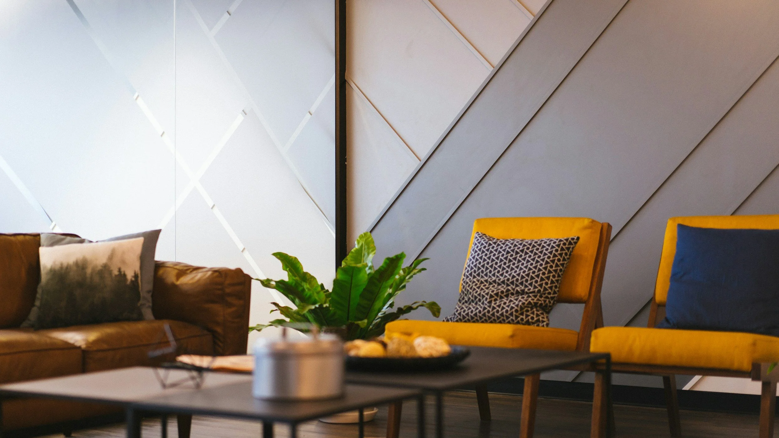 Living room with yellow and blue chairs, a brown sofa with patterned pillows, a small table with snacks, a houseplant, and textured wall decor.
