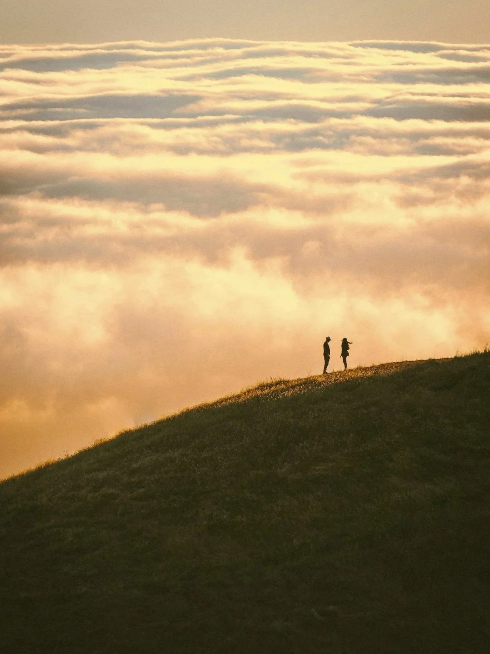 Two people standing on a grassy hill at sunset, silhouetted against a sky with layered clouds illuminated by the setting sun.