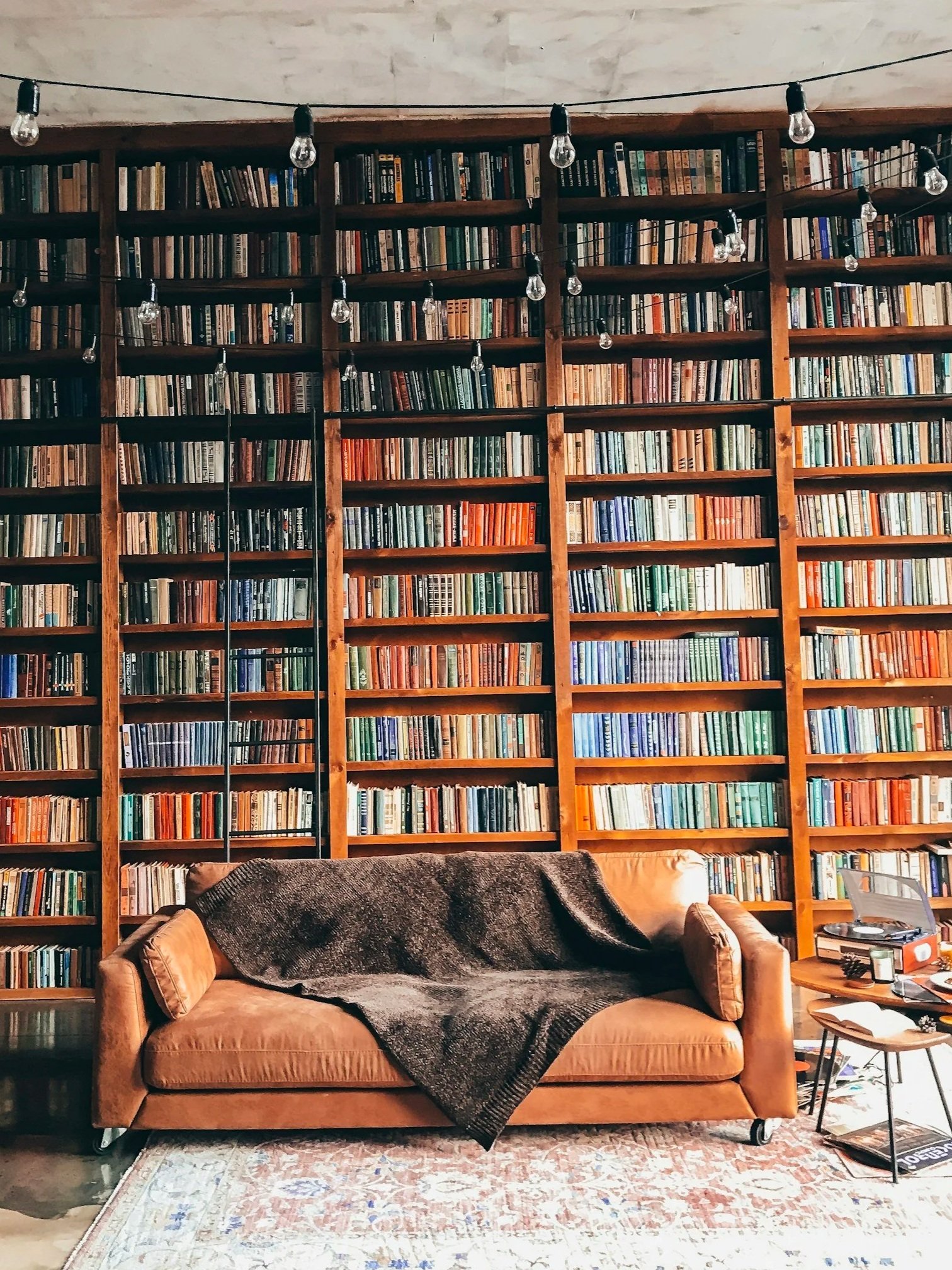 A cozy living room with a large wooden bookshelf filled with books, a tan leather sofa with a dark throw blanket and cushions, a patterned area rug, and a small round side table holding a few magazines, a journal, and a record player with records next to it.