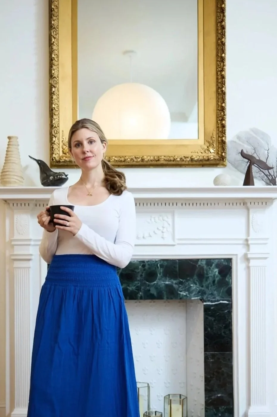 A woman with light brown hair tied back, wearing a white long-sleeve shirt and a blue skirt, stands indoors holding a black mug. She is in front of a white decorated fireplace with a black marble insert, adorned with decorative objects including a black bird-shaped ornament, a beige vase, and a metal sculpture of a bird on a branch. A large ornate gold-framed mirror hangs above the fireplace, reflecting a soft glowing light.