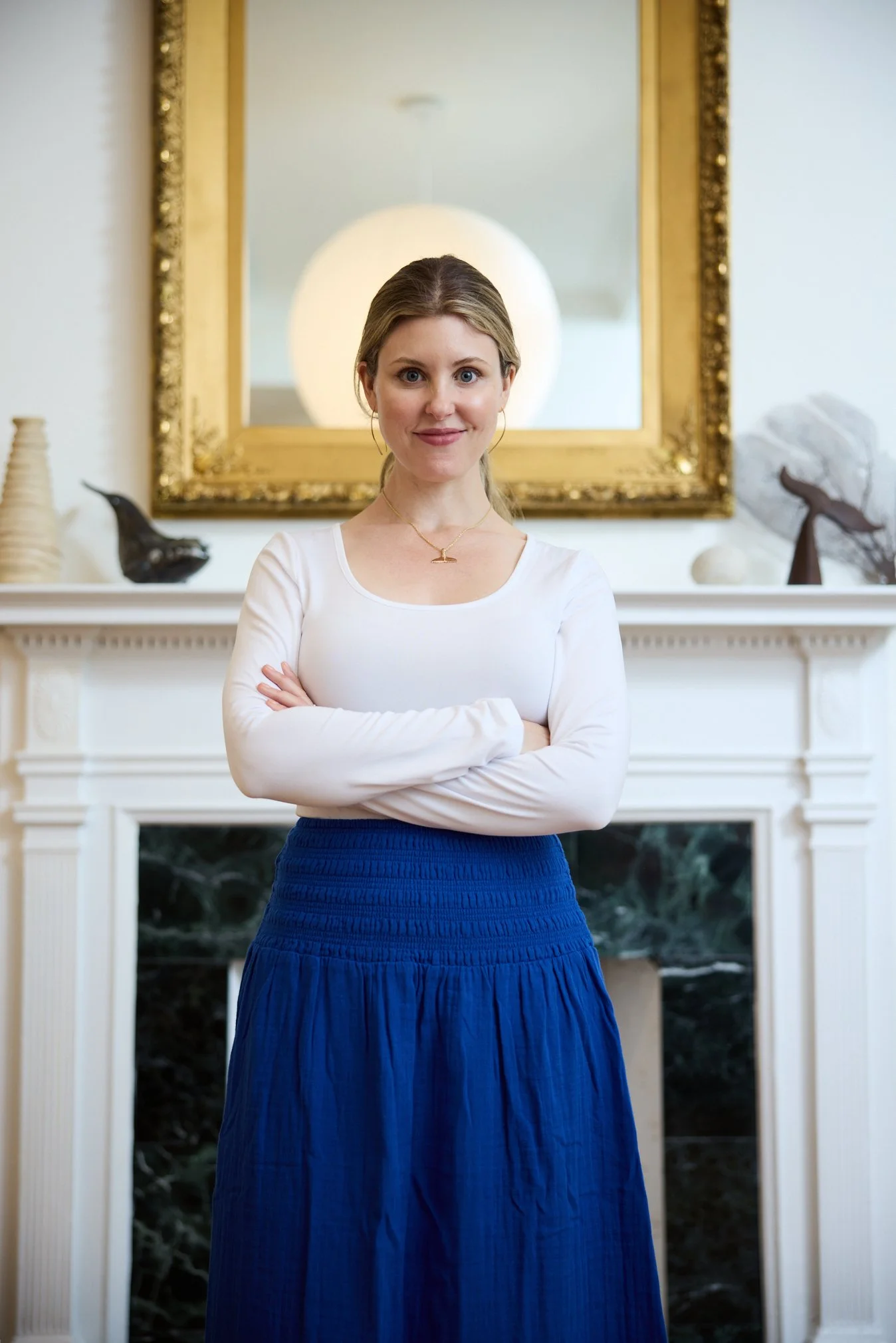 A woman with blonde hair wearing a white long-sleeve top and a blue skirt standing in front of a fireplace with a large mirror and decorative objects.