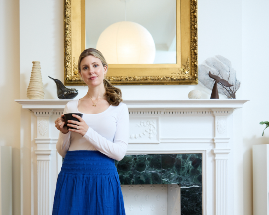 Woman in white top and blue skirt holding a black mug in front of a decorated fireplace with a gold framed mirror above.