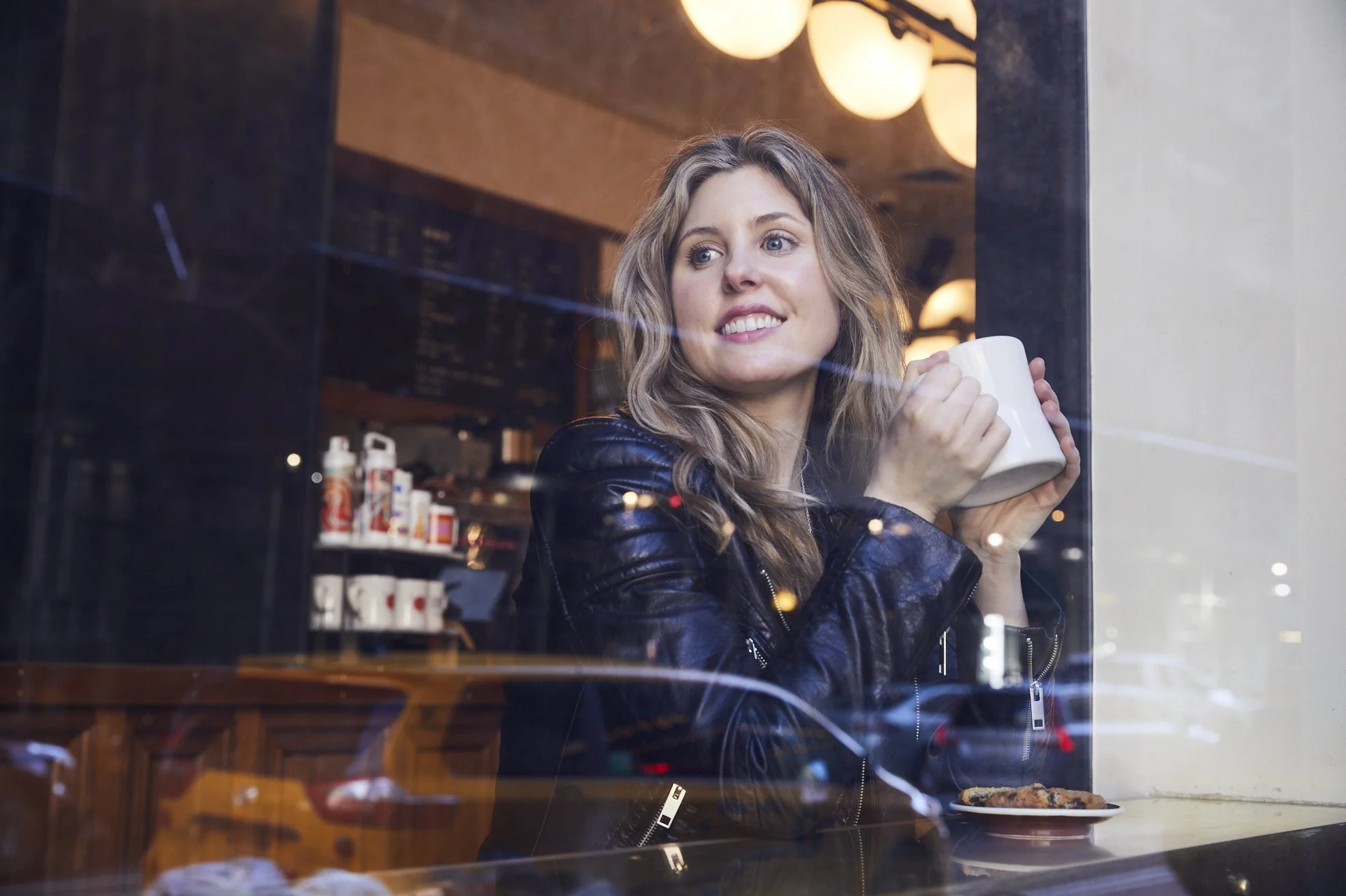 A woman with wavy hair and a leather jacket sitting inside a coffee shop, looking through the window and holding a white mug. The shop interior has warm lighting, a menu board, and some bottles on the shelf.