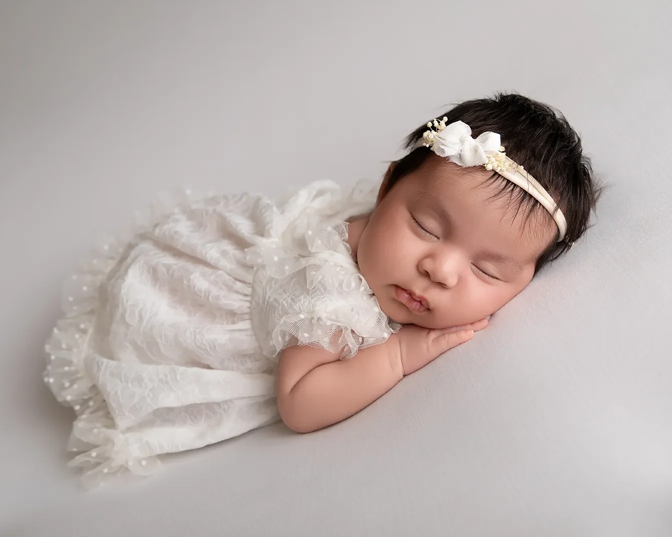  Newborn baby girl photographed in a Glendale California studio with soft neutral tones and dressed in a white lace dress. 