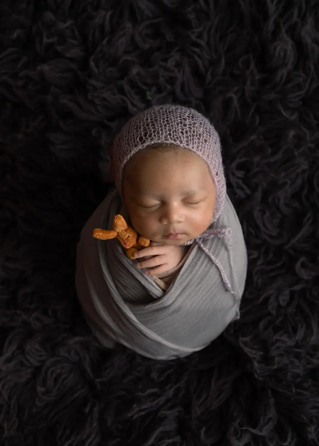  newborn boy on dark flokati in potato sack pose holding a stuffie. 