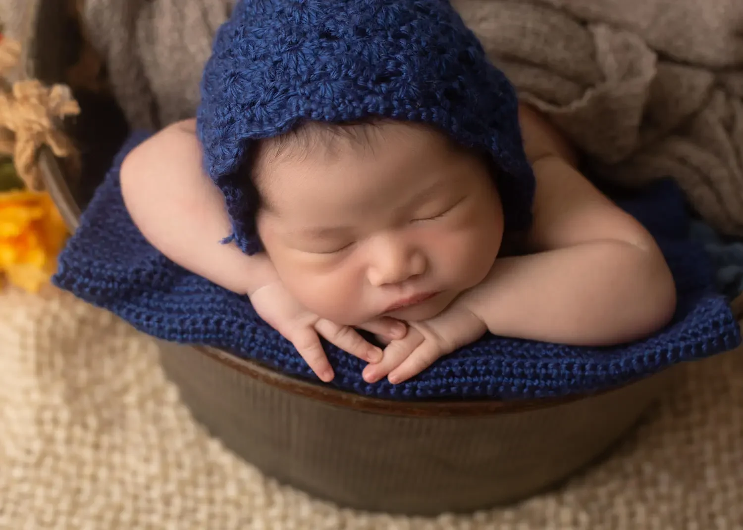  newborn boy in metal bucket with chin on hands. Close up. Wearing a knitted blue bonnet. 