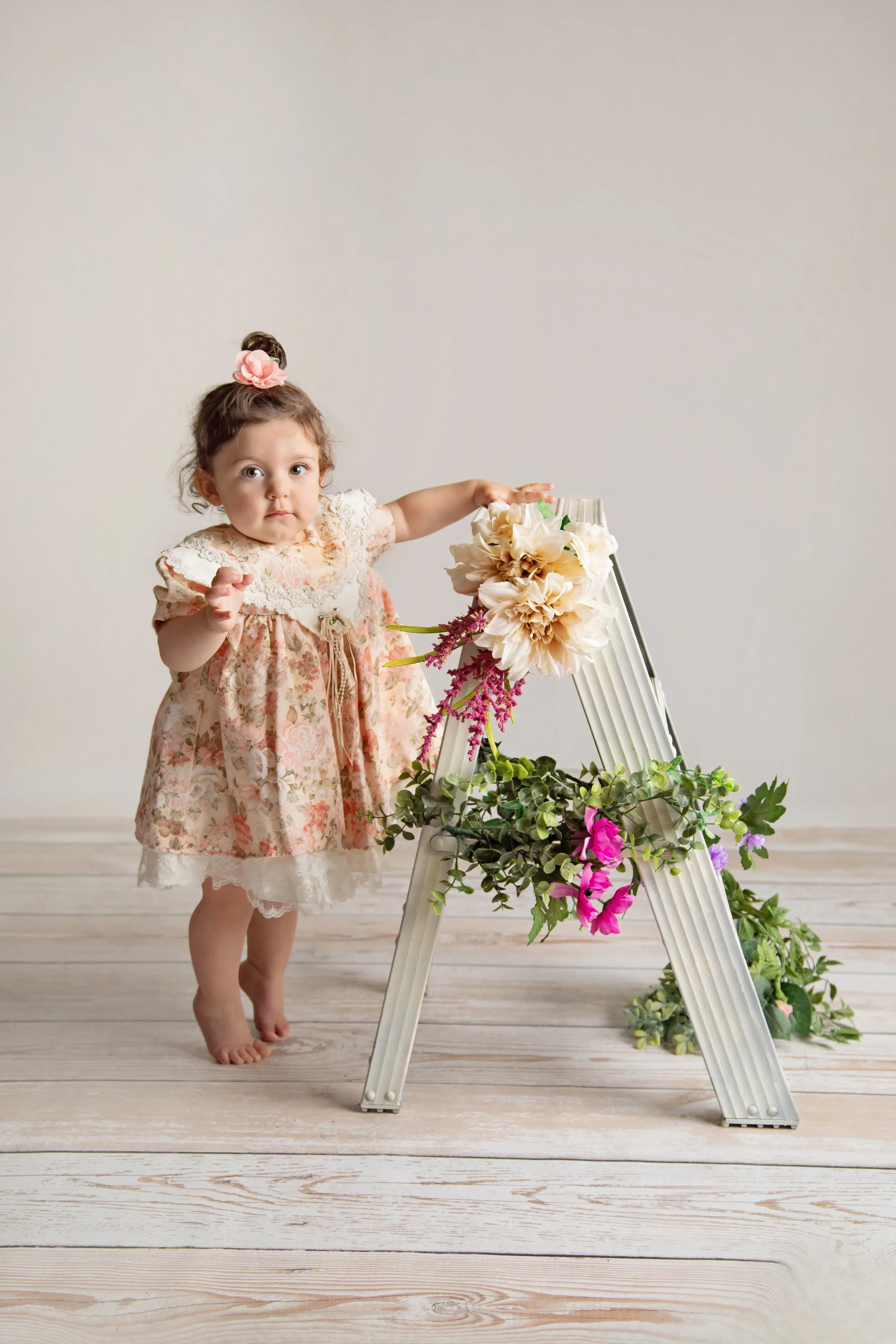 A young girl in a floral dress with a pink hair accessory standing barefoot on a wooden floor, touching a ladder decorated with flowers.