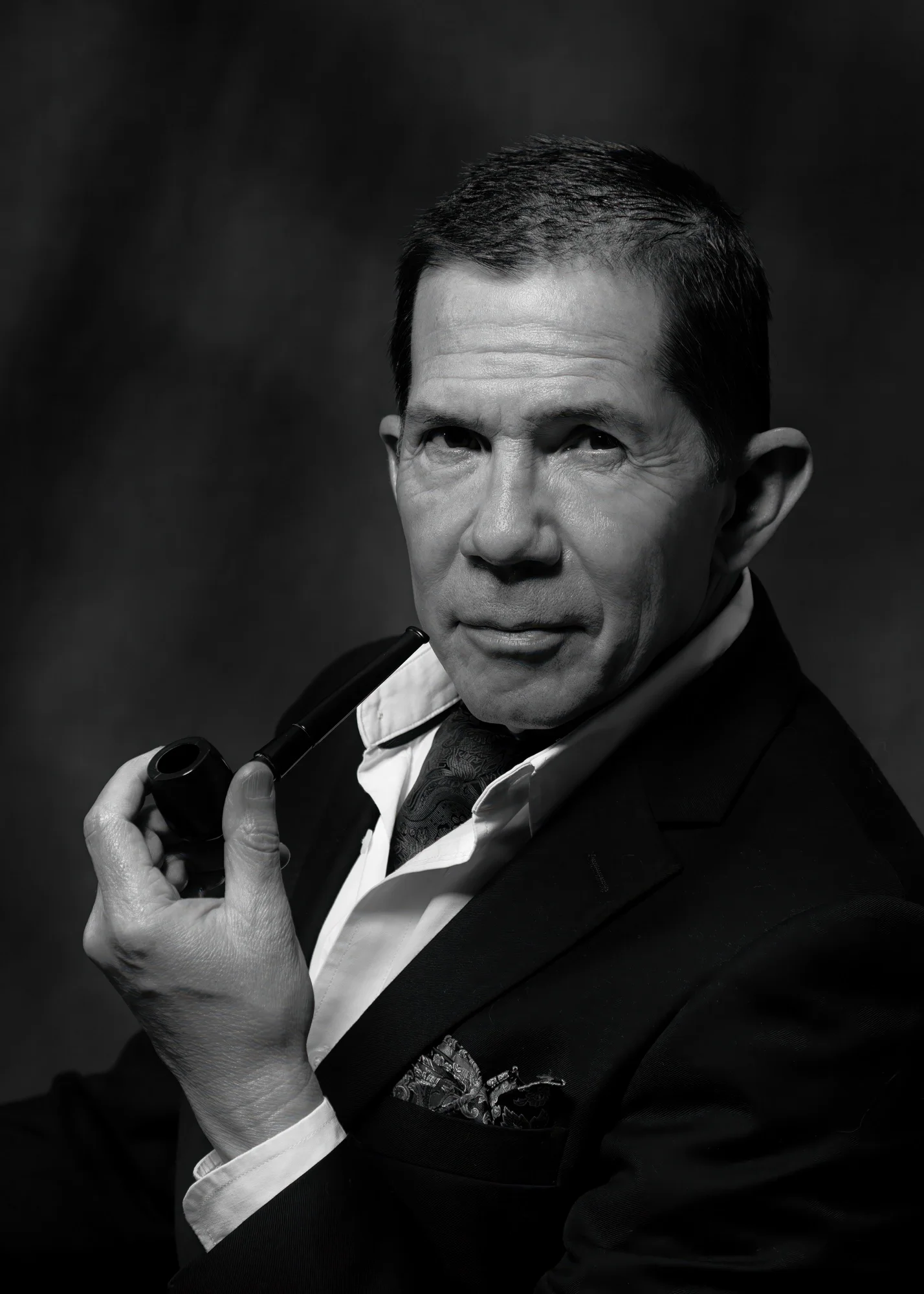 Dramatic black-and-white theatrical headshot of a mature male actor in a suit, posed with a pipe against a dark studio background. Directional lighting, deep shadows, and classic styling create a distinctive character portrait suitable for acting por