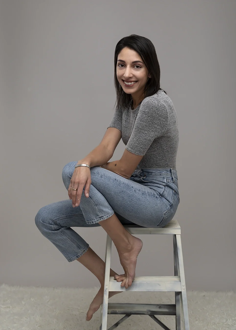 Lifestyle professional portrait of a woman seated casually in a studio against a neutral gray background. The relaxed pose and contemporary styling create an approachable personal branding image suitable for professional websites, social media, creat