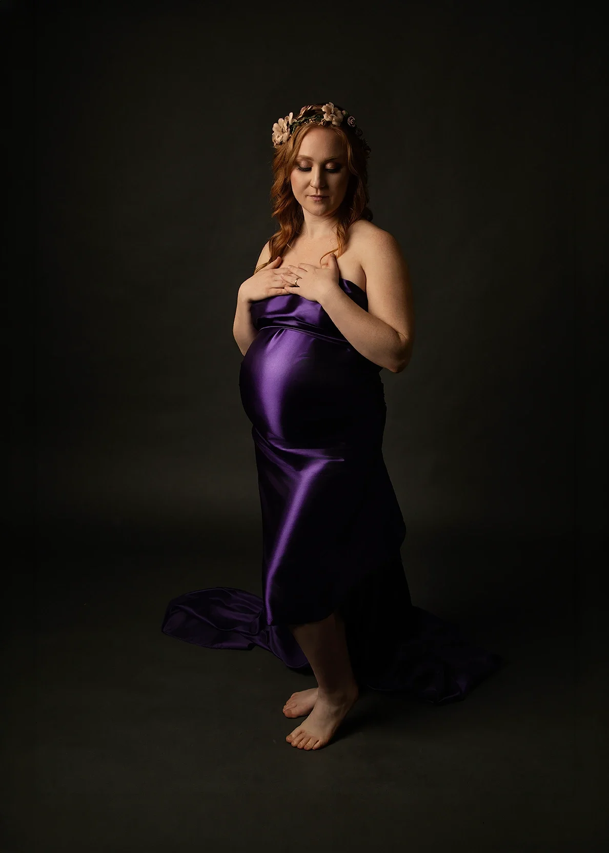 Pregnant woman in a fitted purple maternity gown and floral headpiece posed in a dark fine-art studio portrait, photographed late in pregnancy.