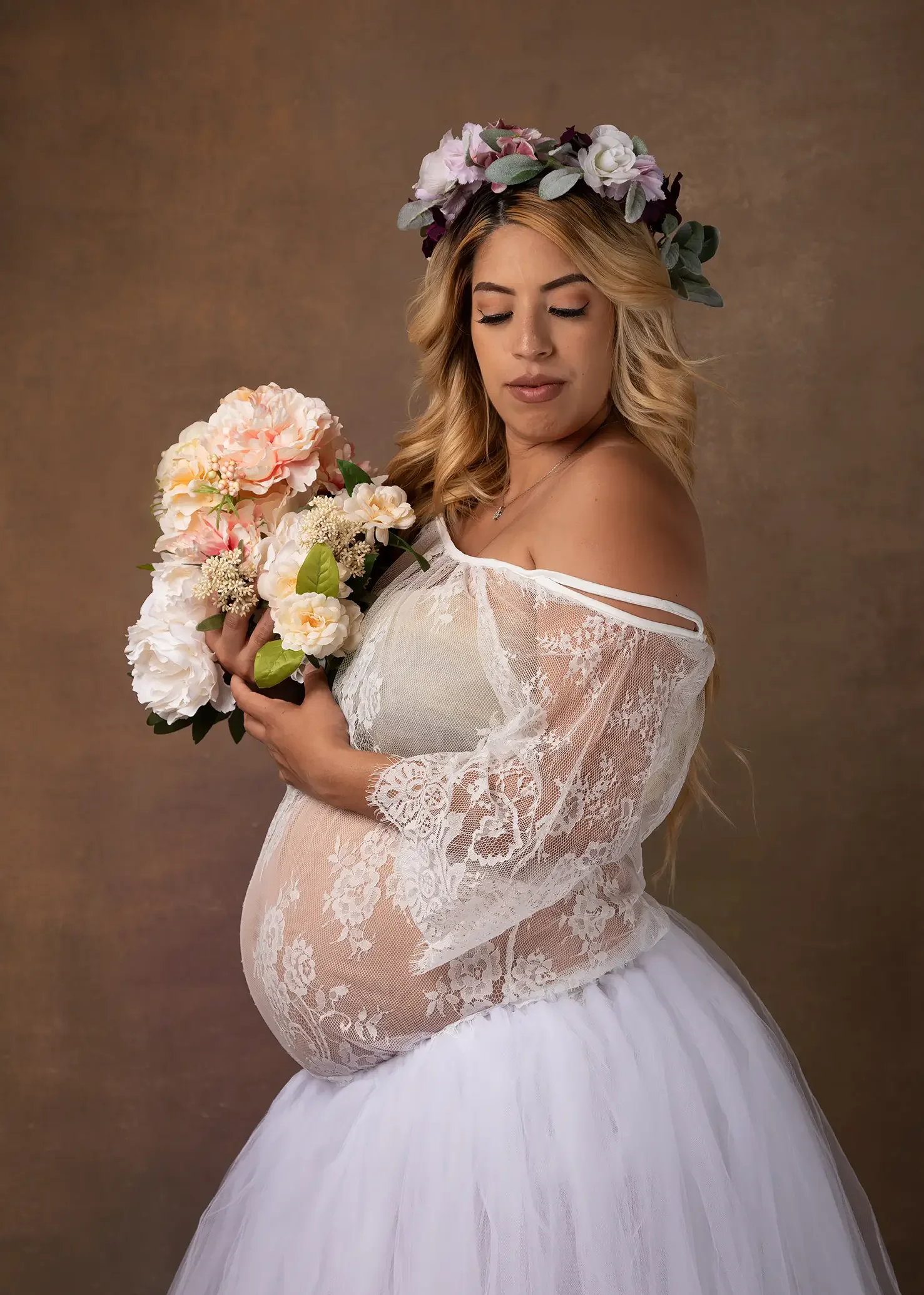 Pregnant woman in a white lace maternity gown and floral crown holding flowers during a studio maternity photography session in Los Angeles.