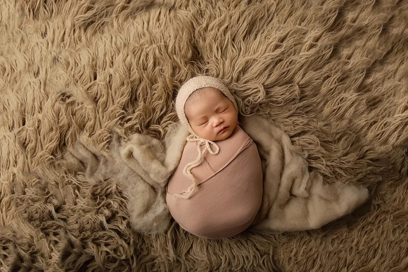 Sleeping newborn baby wrapped in a soft blush wrap and wearing a cream knit bonnet, posed on a textured neutral flokati blanket during a studio newborn photography session in Los Angeles. Timeless wrapped newborn portrait with soft earth tones.
