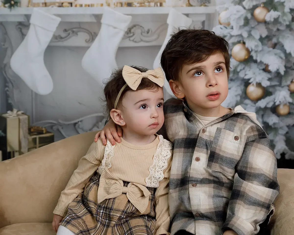 Brother and baby sister sharing a quiet holiday moment during a studio family Christmas photo session in Glendale, Los Angeles with a decorated fireplace and Christmas tree.