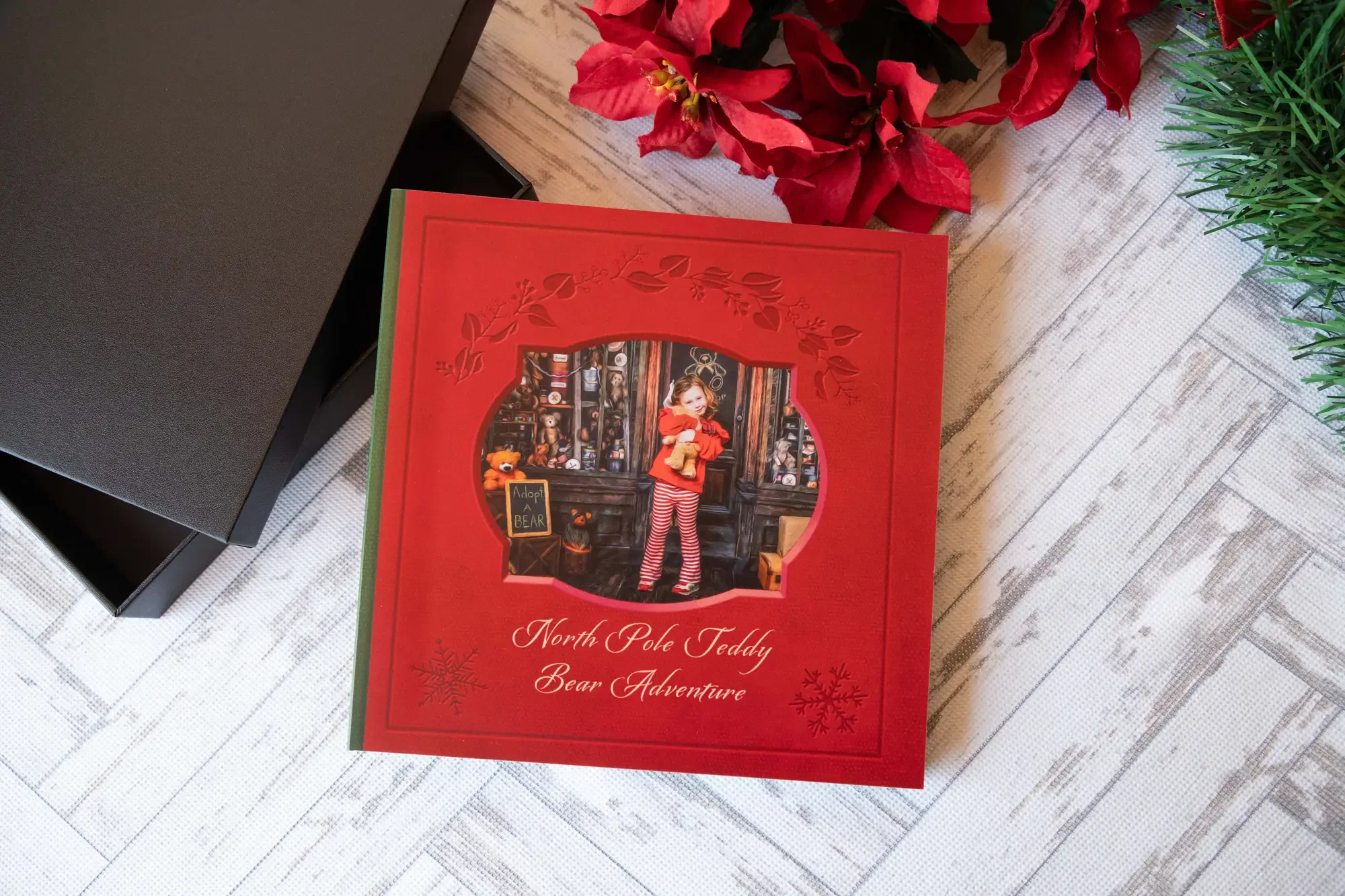 Red North Pole Teddy Bear Adventure storybook featuring a child holding a teddy bear in a Christmas workshop scene, displayed beside a black keepsake box and holiday greenery.