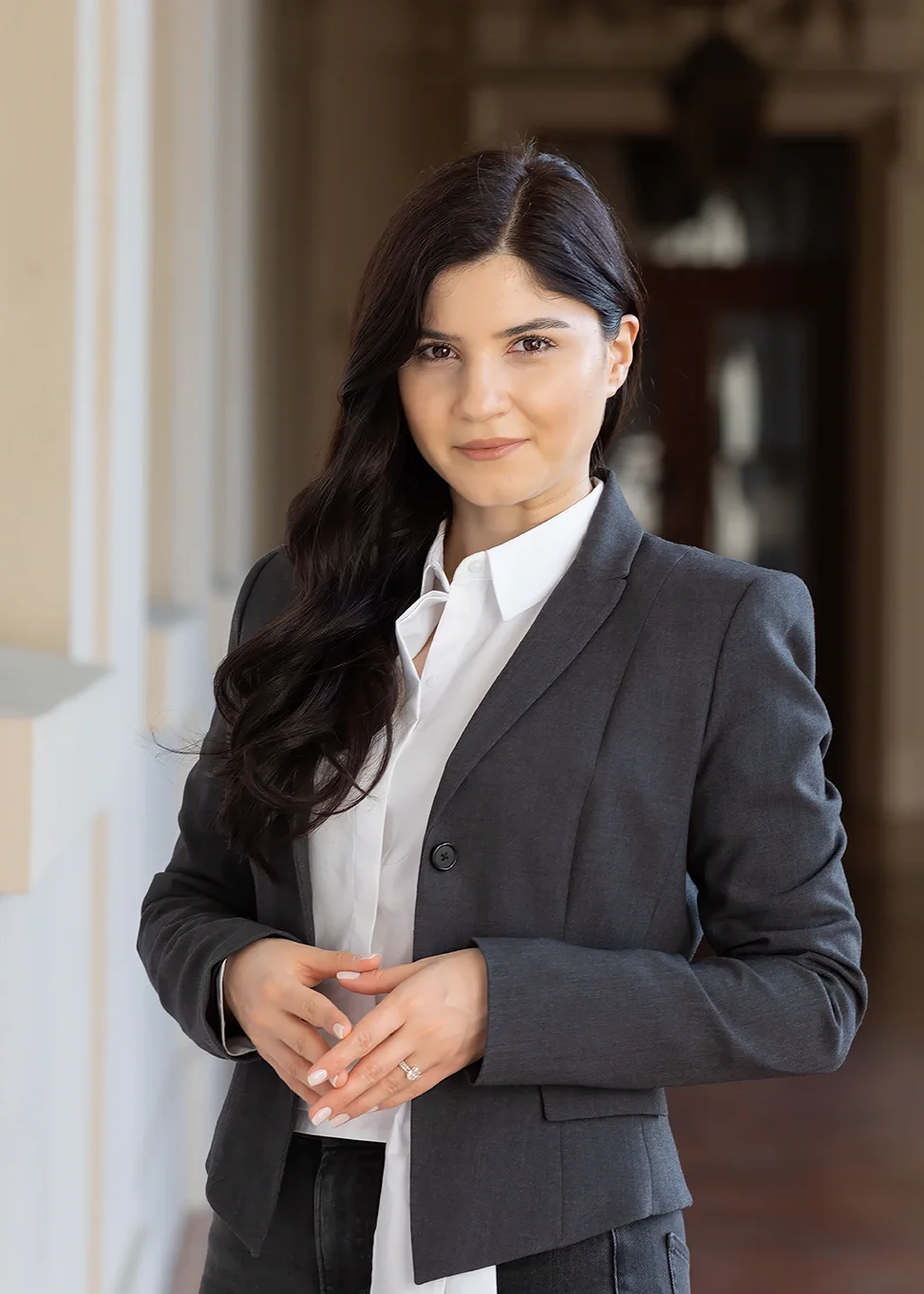 Professional corporate portrait of a woman in a gray blazer and white blouse photographed in a softly lit outdoor setting. The polished styling and environmental background create a confident, approachable image suitable for executive profiles, compa