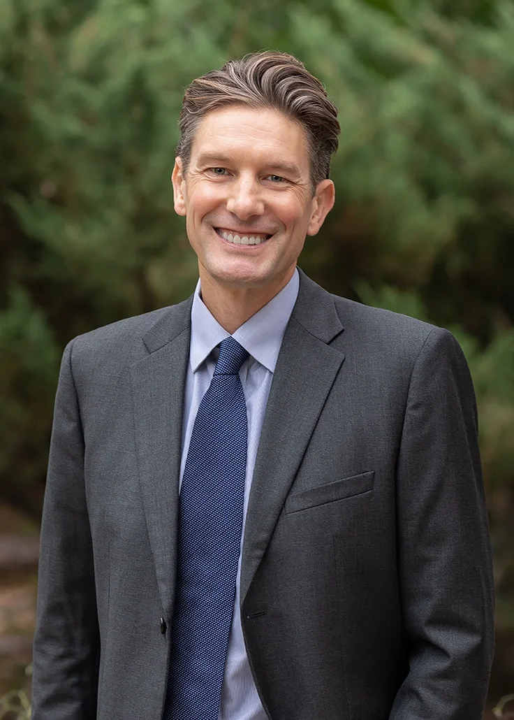 Professional outdoor headshot of a man in a suit and tie photographed against a softly blurred natural background. The relaxed expression and polished styling create an approachable professional portrait suitable for physicians, medical professionals