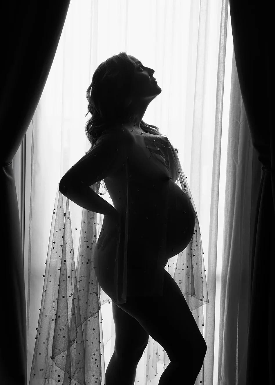 Black-and-white silhouette maternity portrait of a pregnant woman posed in sheer draped fabric against a bright window, emphasizing the shape of her baby bump.