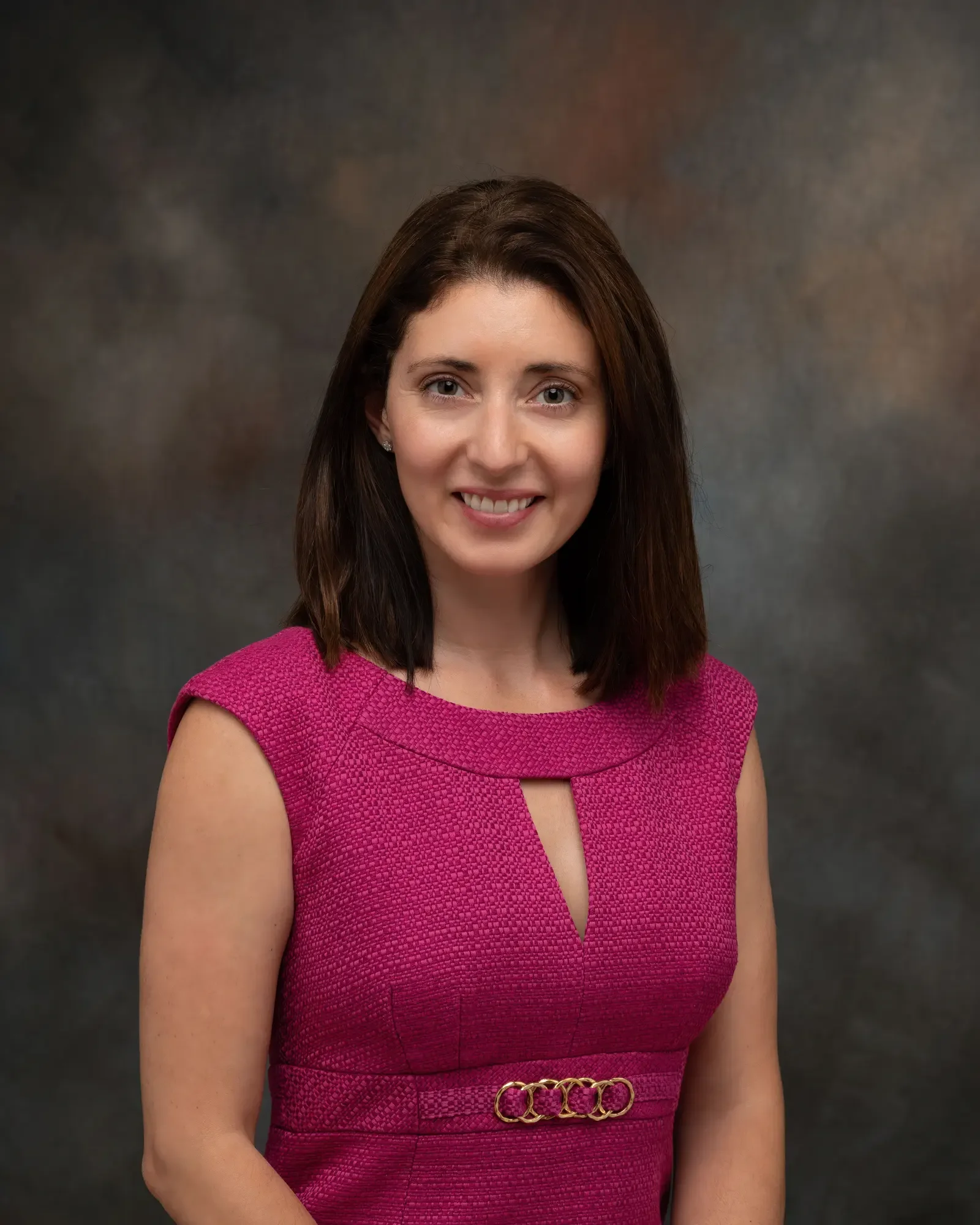 
Professional corporate headshot of a woman in a magenta dress photographed against a textured gray studio background. The classic portrait lighting and simple composition create a polished, approachable image suitable for executive profiles, company