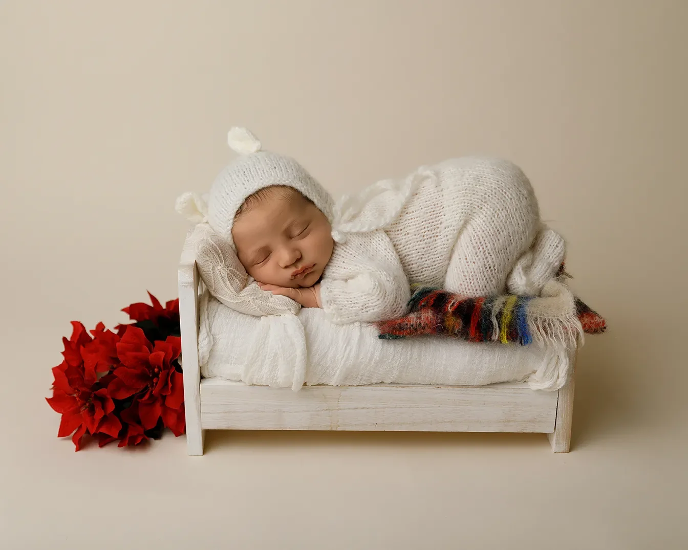 Sleeping newborn wearing a white knit outfit and bonnet posed on a miniature white bed with a plaid blanket and red poinsettias during a studio newborn photography session. in los angeles.