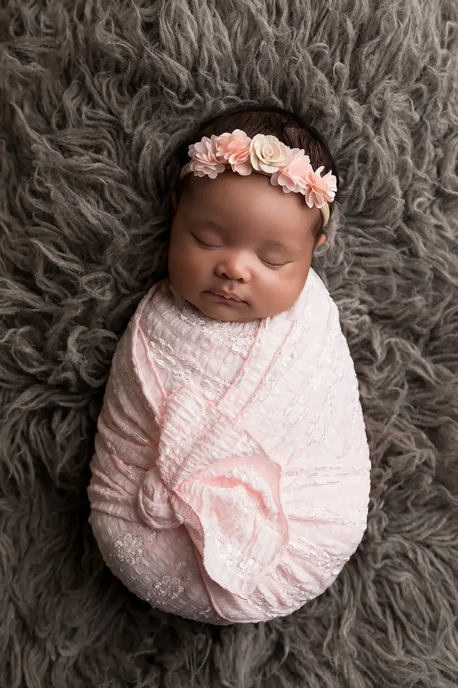 Sleeping newborn baby girl wrapped in a blush pink swaddle and wearing a floral headband on a soft gray fur blanket during a professional studio newborn photography session in Los Angeles.