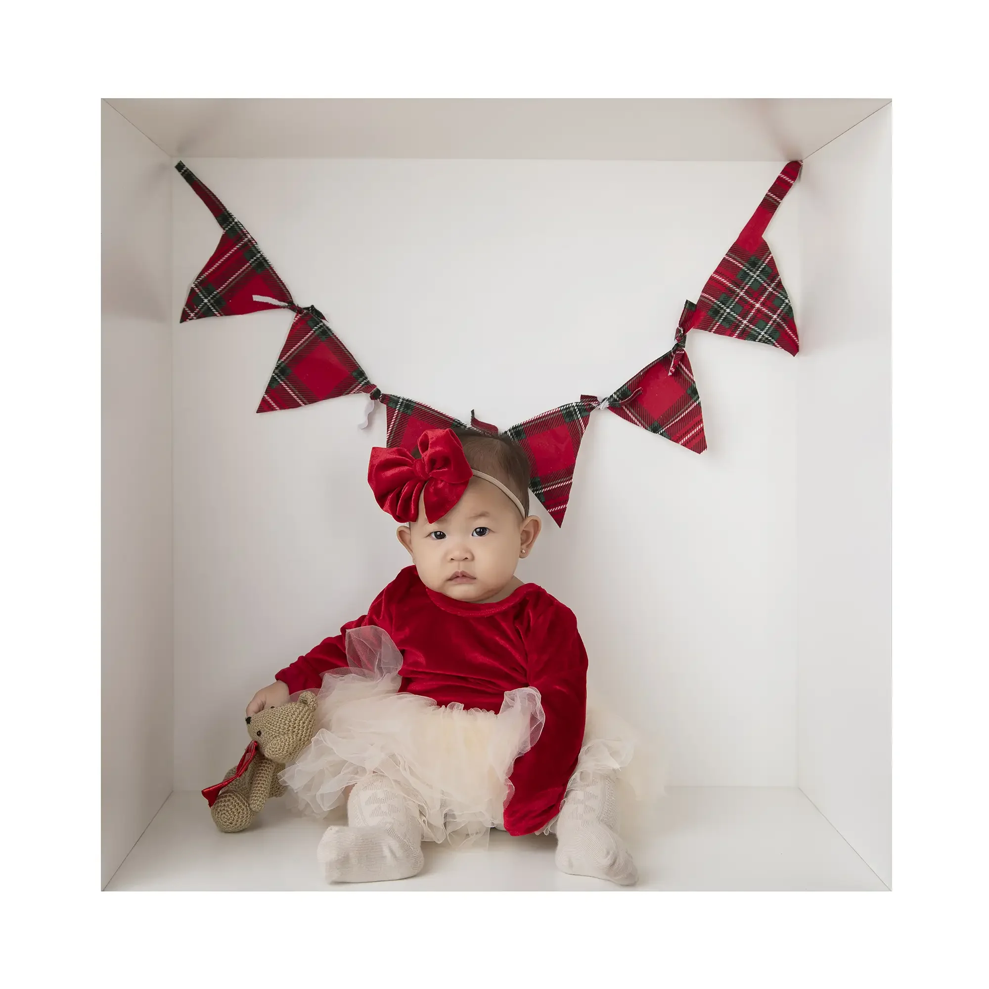 Baby girl wearing a red velvet holiday dress and white tutu sitting inside a Baby in a Box Christmas portrait with festive plaid bunting during a first Christmas photo session in Glendale, Los Angeles.