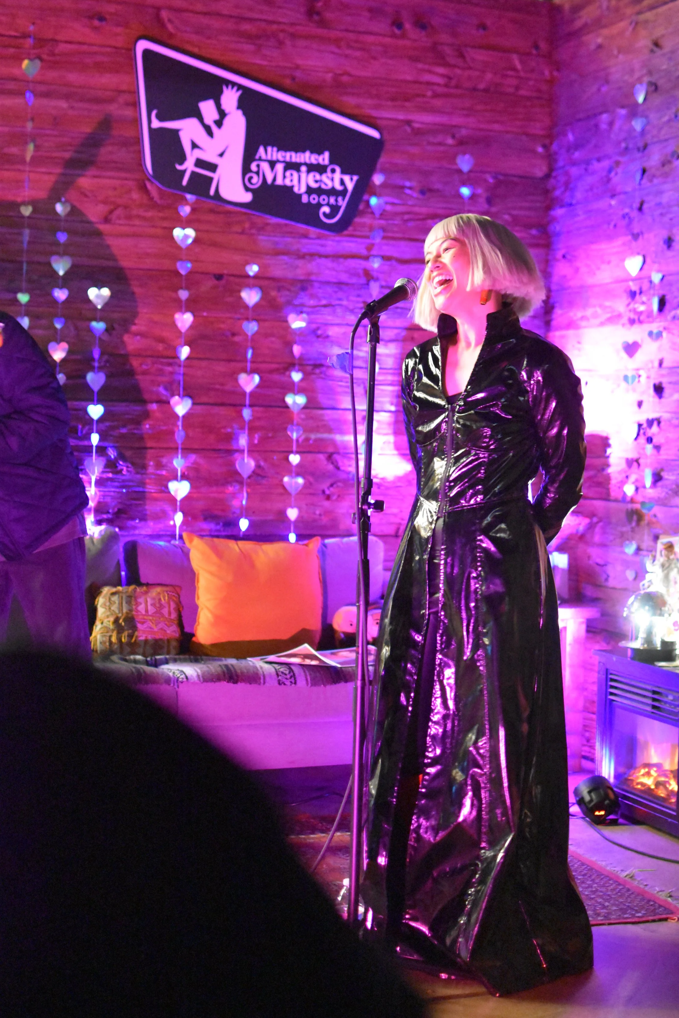 promqueen (musician) standing and singing into a microphone. they are wearing a long black faux leather coat.