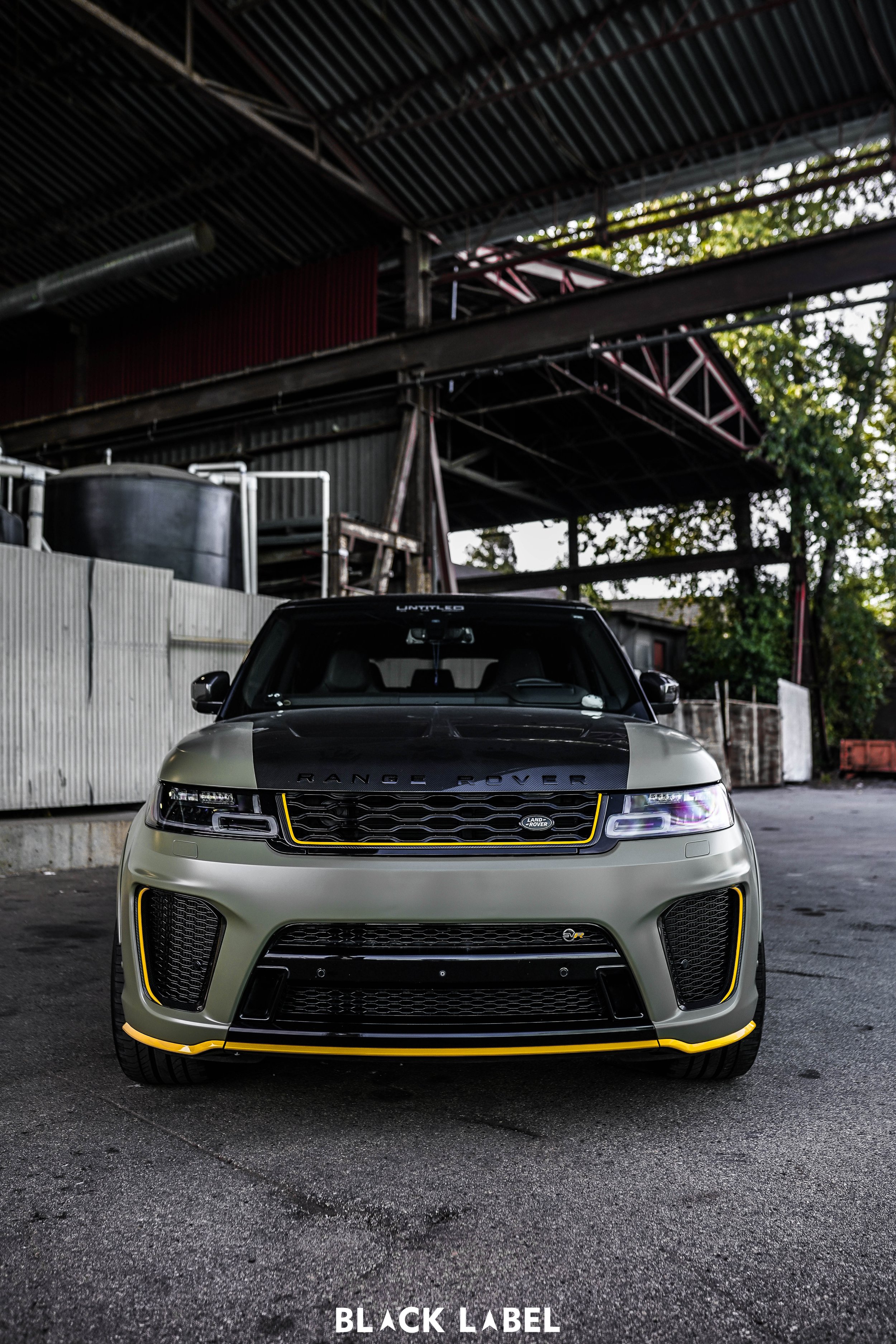 Front view of a two-tone black and gray Range Rover SUV with yellow accents, parked outdoors under a metal structure with trees in the background.