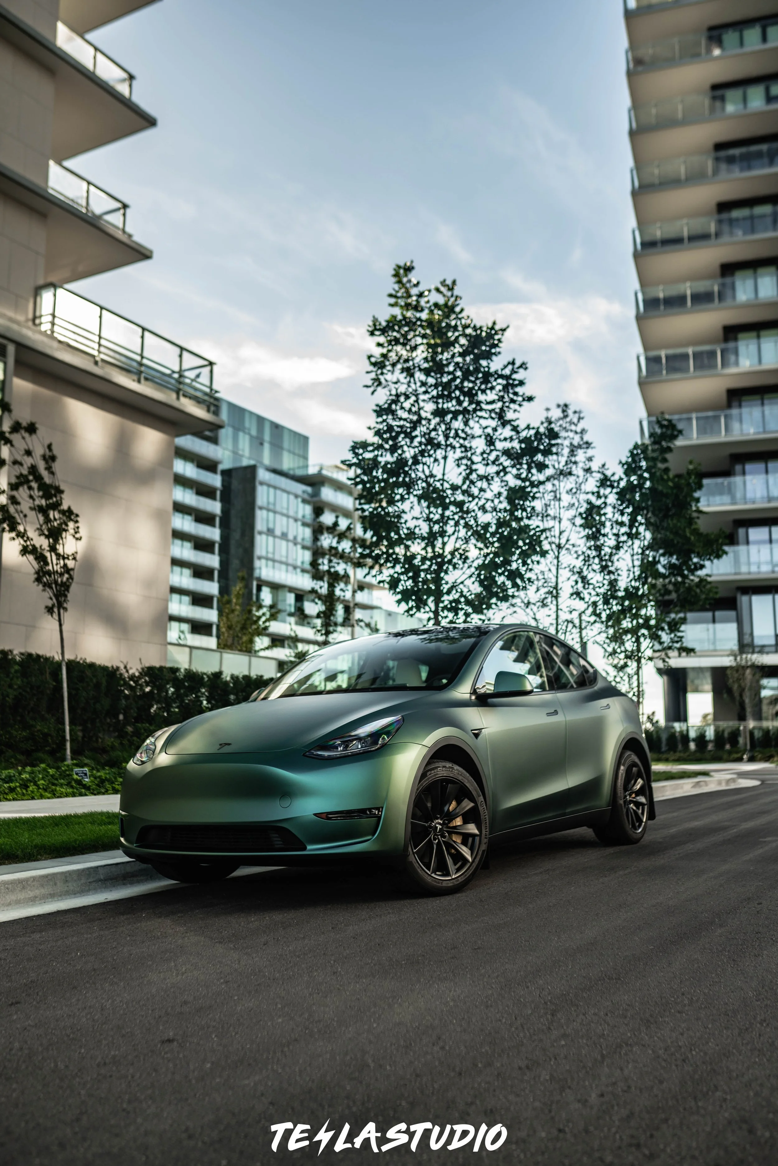 A green Tesla Model Y parked on a residential street with modern apartment buildings and trees in the background.