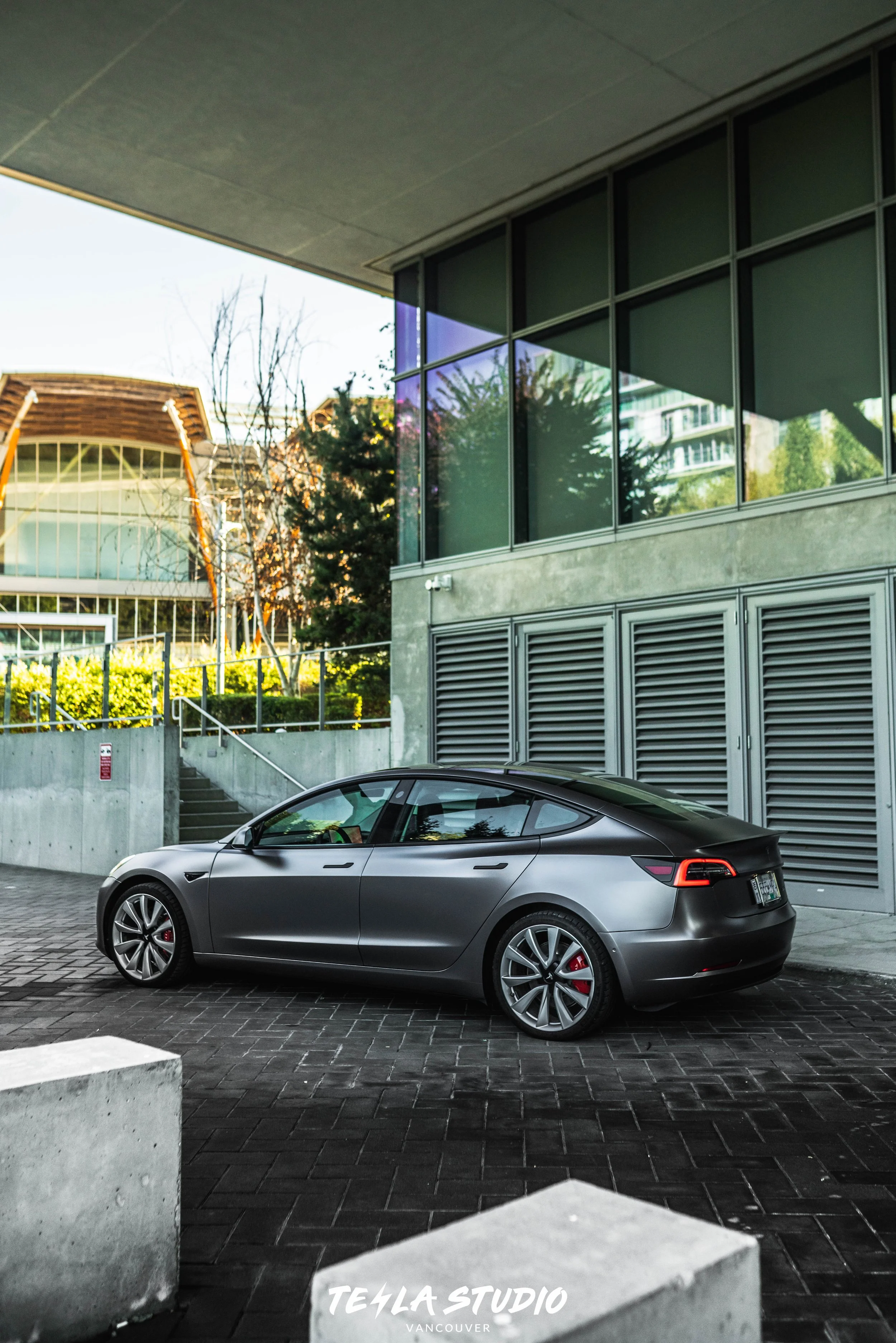 A sleek, metallic gray Tesla Model S parked outside a modern building with large glass windows, surrounded by concrete and landscaped greenery.