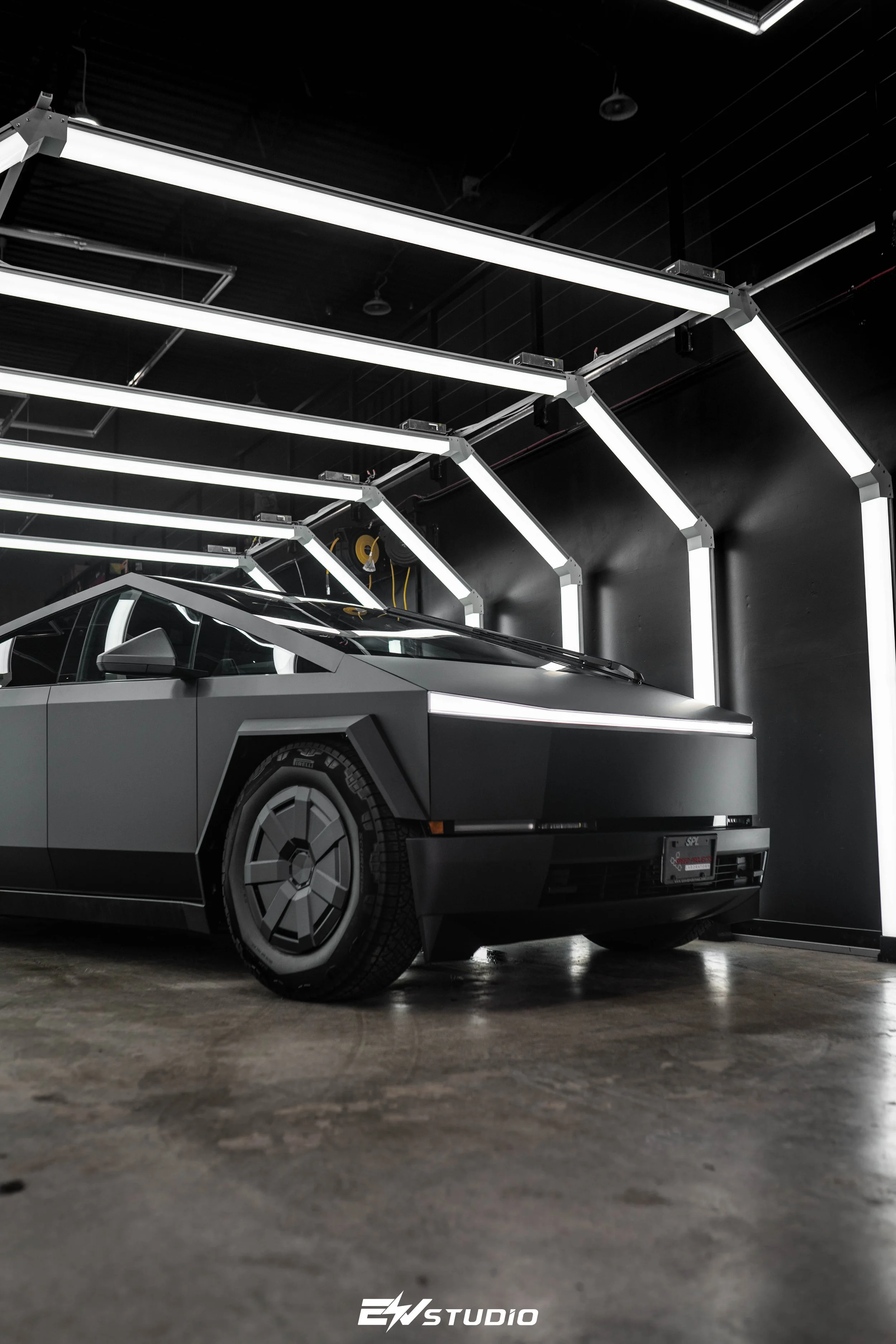 A futuristic, matte black car with angular design parked indoors under bright white neon lighting arranged in geometric patterns.