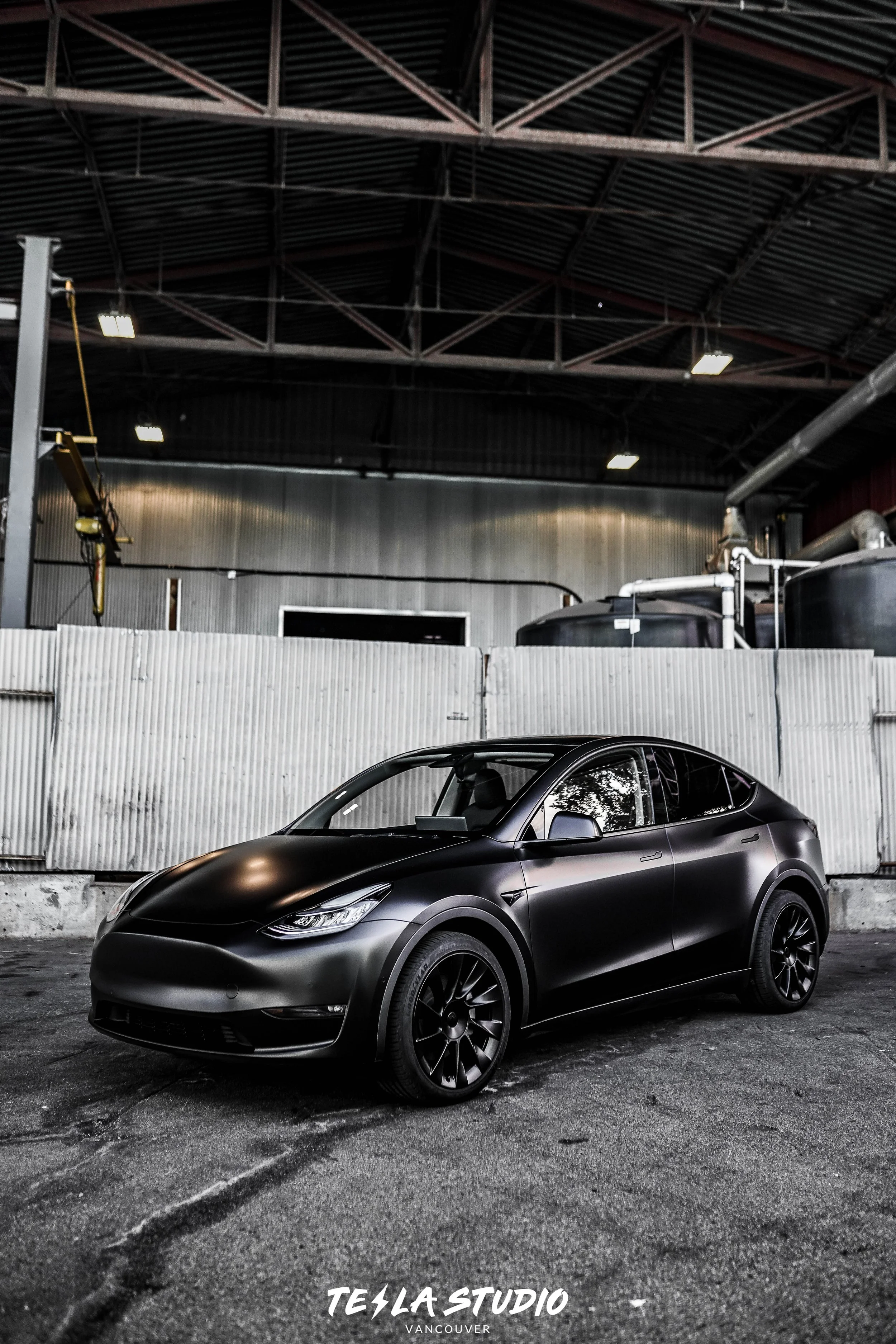 A sleek black Tesla Model Y parked inside an industrial warehouse with a metal roof and walls, and some large equipment in the background.