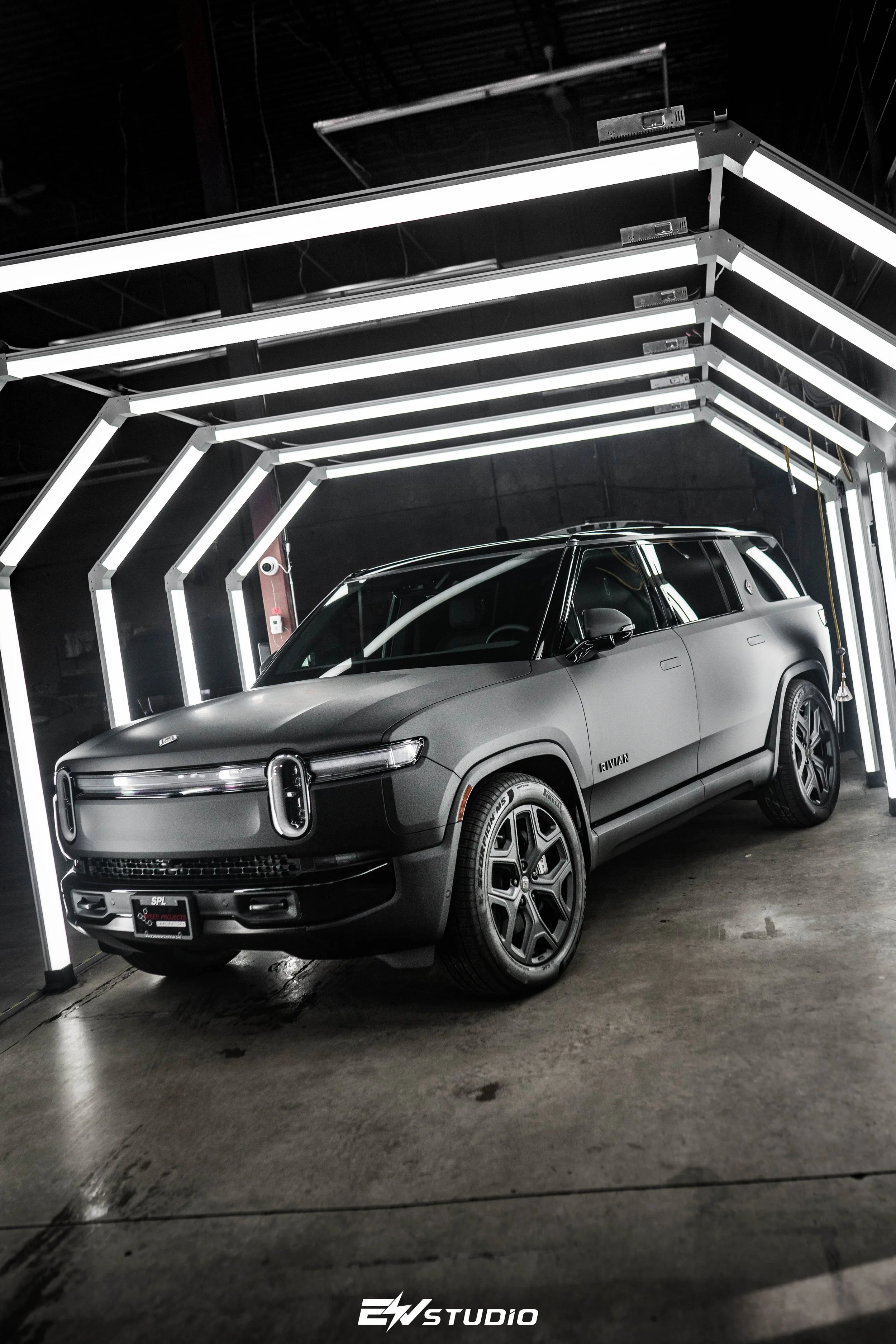 A matte black Rivian R1T electric pickup truck in a studio with LED strip lighting overhead, reflecting on the polished concrete floor.