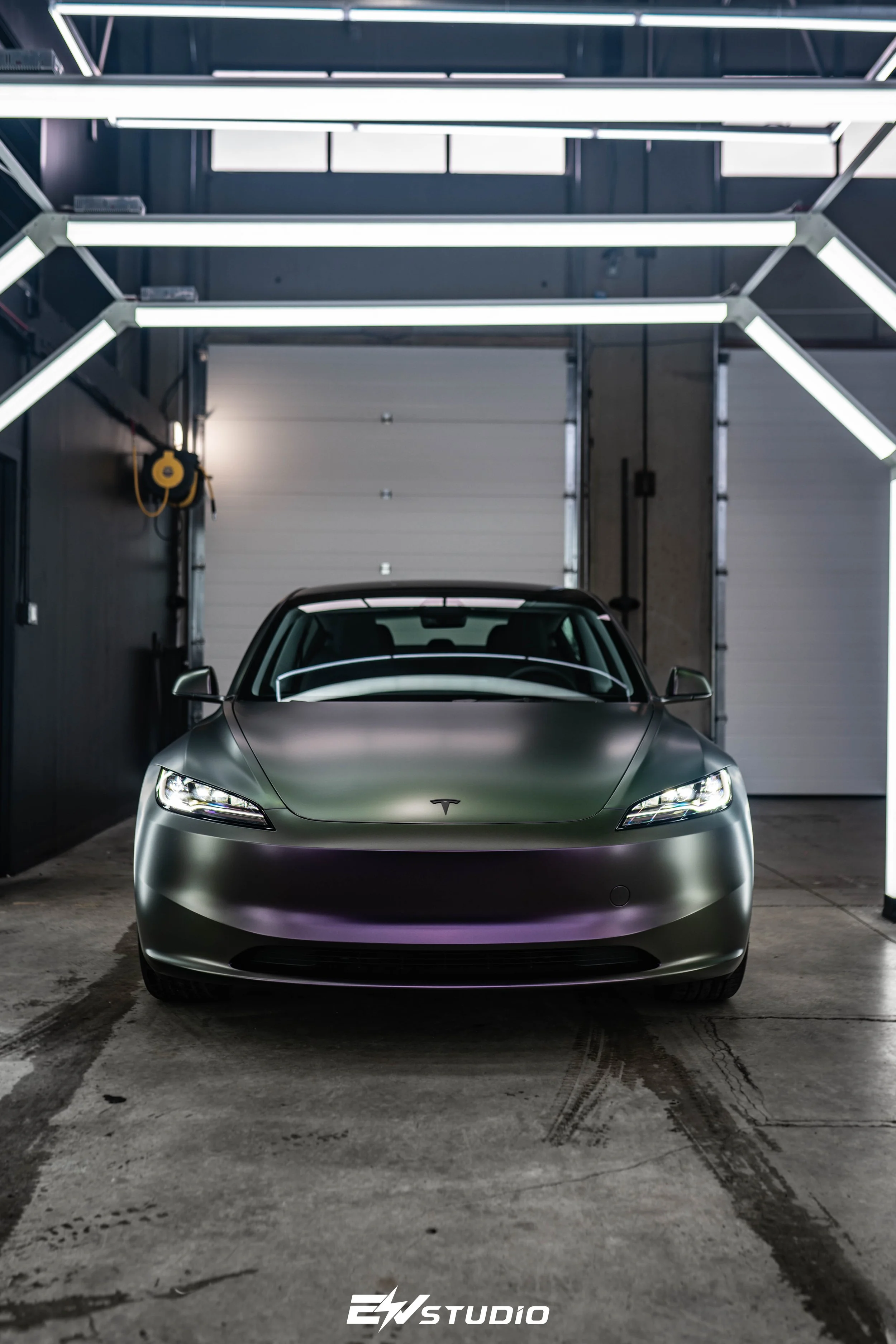 A matte gray Tesla Model 3 parked inside a garage with modern LED lighting overhead